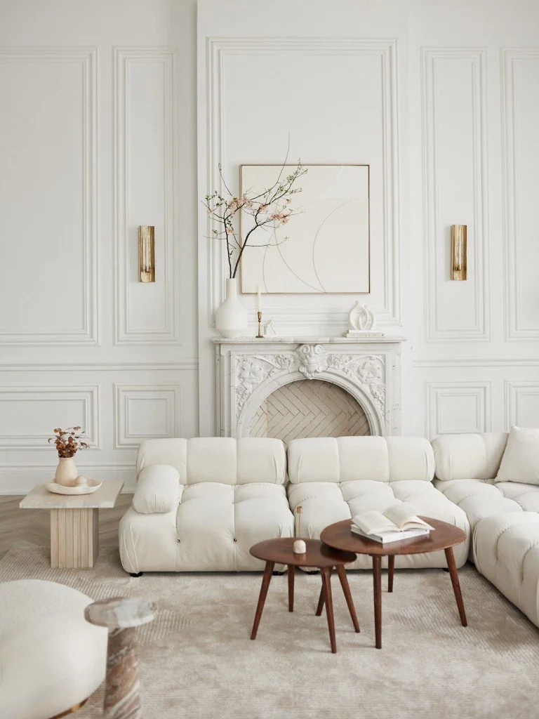 Elegant living room with white paneled walls, a white tufted sofa, a marble fireplace, and minimalist decor including a large white vase with branches. Two small wooden tables sit in front of the sofa, with an open book and a small candle on one. A small side table with a decorative object and a side wall sconce are also visible.