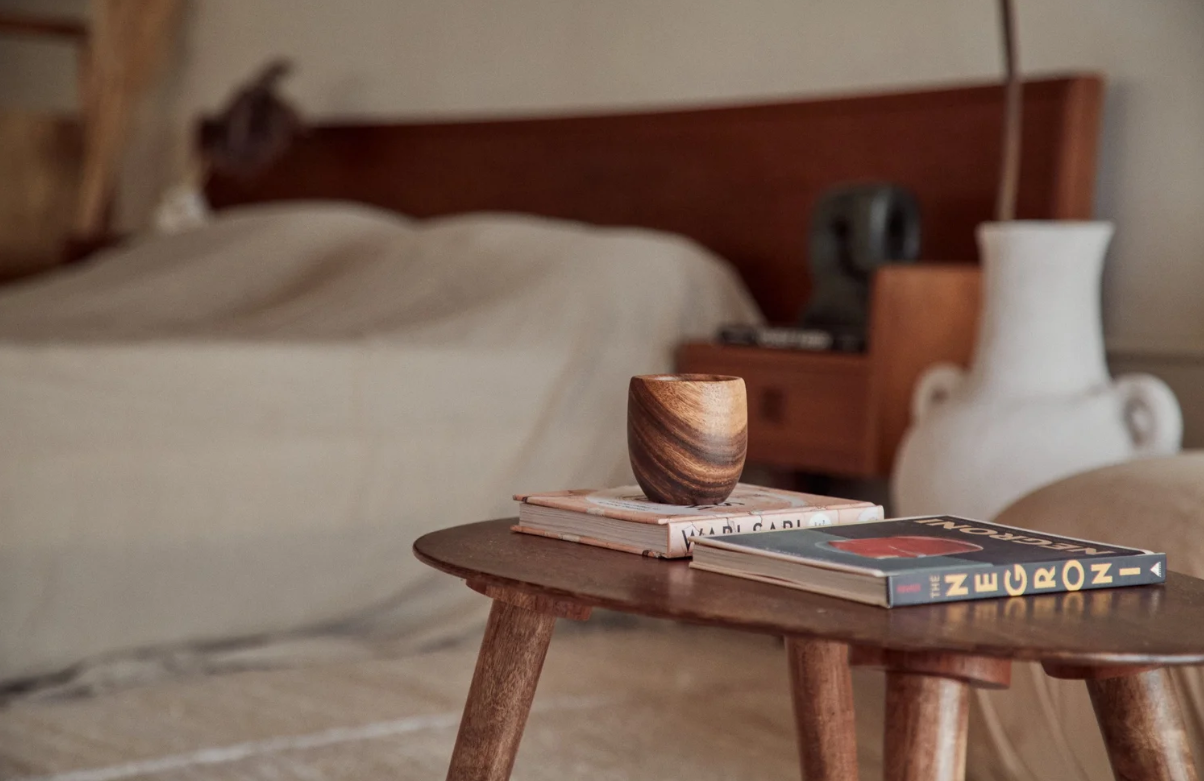 Close-up of a small wooden table with a wooden cup, two magazines, and a book on top, in a cozy bedroom setting with a bed, nightstand, and decorative vase in the background.