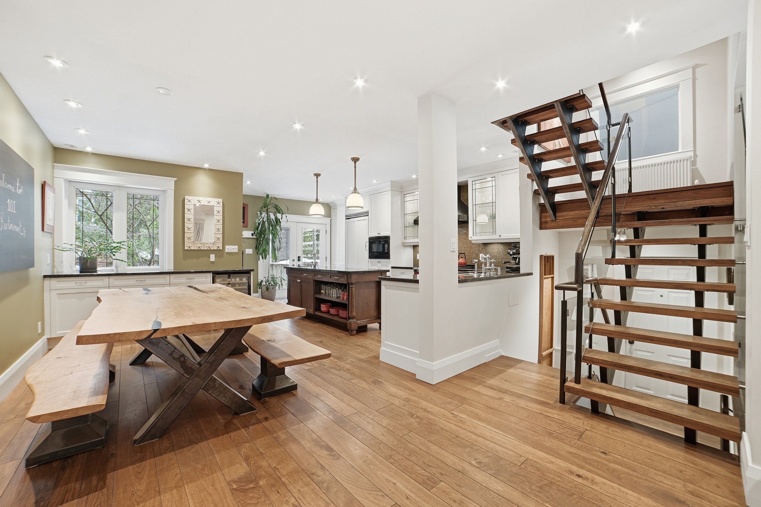 Open-concept kitchen and dining area with wooden flooring, a large wooden dining table with benches, white cabinetry, black countertops, and a staircase with wooden treads and black metal railing.