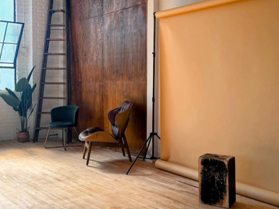 Indoor photography studio with a beige backdrop, three chairs of different styles, a ladder, a potted plant, and a black box.