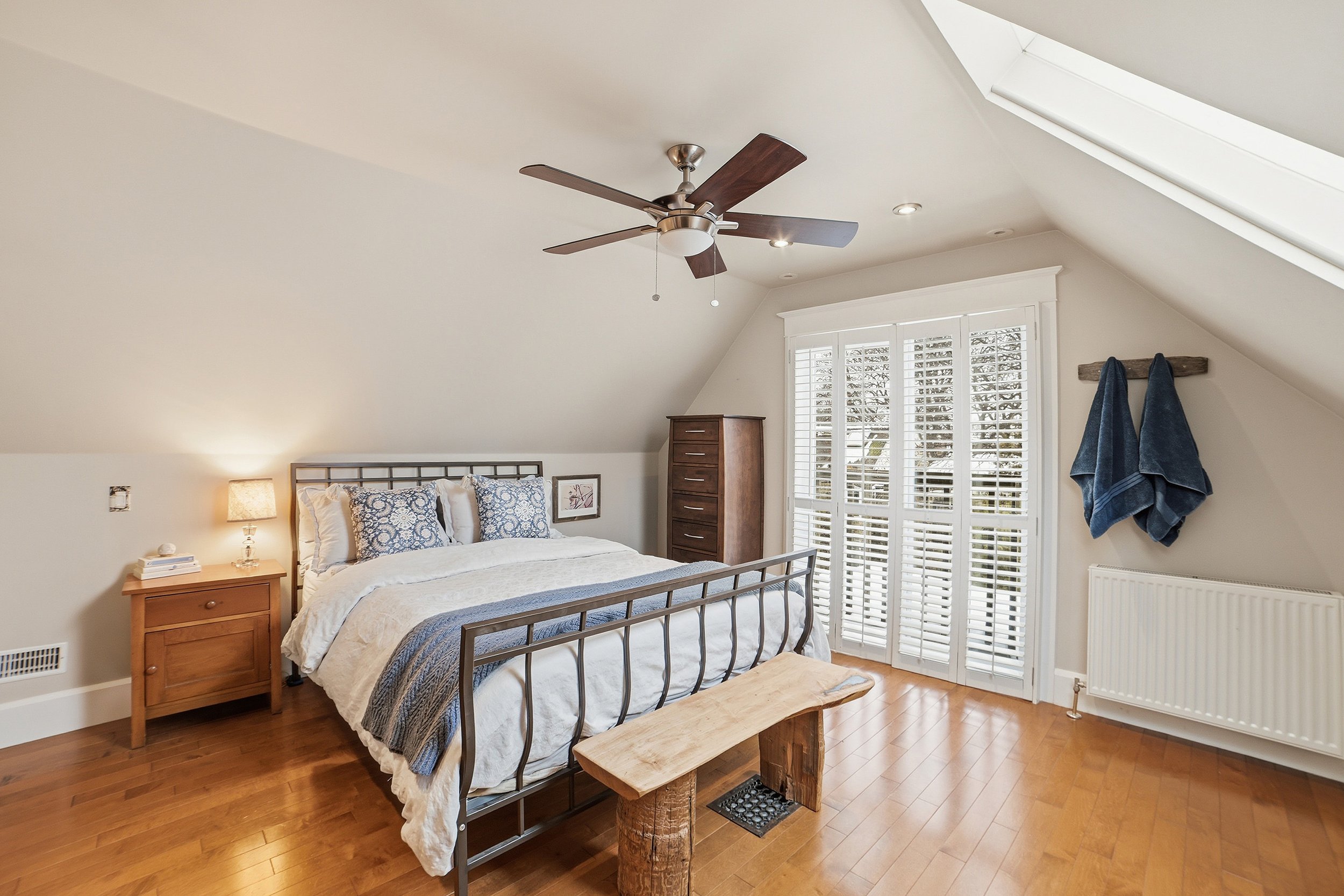 A bedroom with a steel bed frame, white bedding, blue patterned pillows, a wooden nightstand with a lamp, a tall wooden chest of drawers, a bench at the foot of the bed, and shutters on the window, with sloped ceiling and skylight.