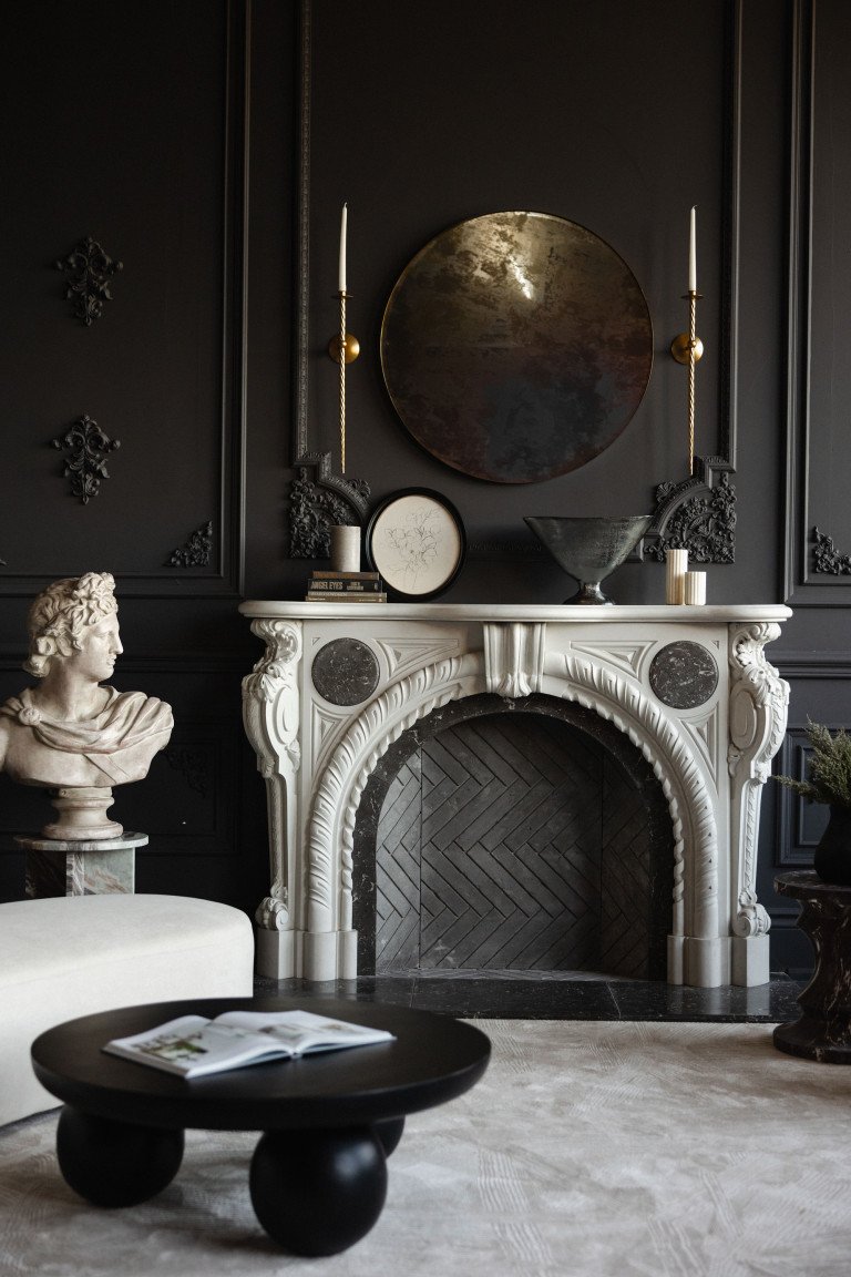 A lavish living room with black walls and ornate decor, featuring a white marble fireplace with black accents, a classical bust sculpture on a small marble pedestal, a black side table with an open book, and a black vase with green foliage. The fireplace mantel is decorated with candles, books, and art pieces, with gold and black candle sconces on the wall and a large round mirror above the fireplace.