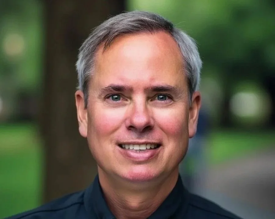 A middle-aged man with short gray hair, blue eyes, and a friendly smile, wearing a dark shirt, outdoors with blurred green trees in the background.