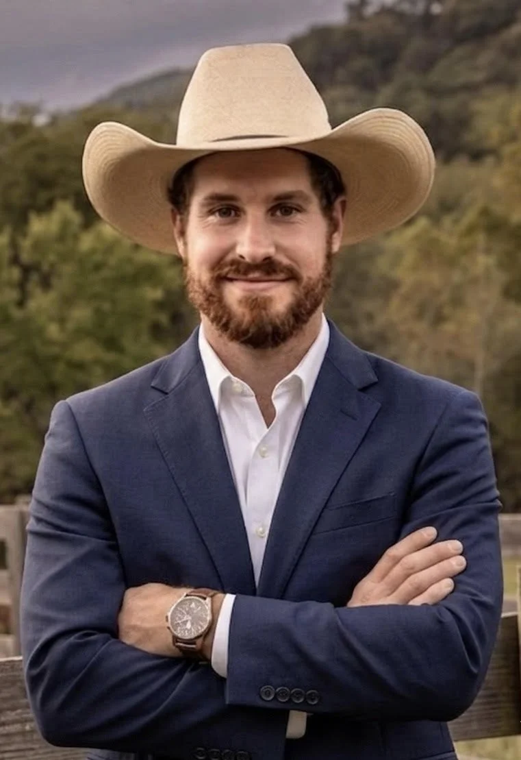 A man with a beard wearing a cowboy hat, a navy blue suit, white shirt, and a watch, standing outdoors with a scenic background of trees and hills.