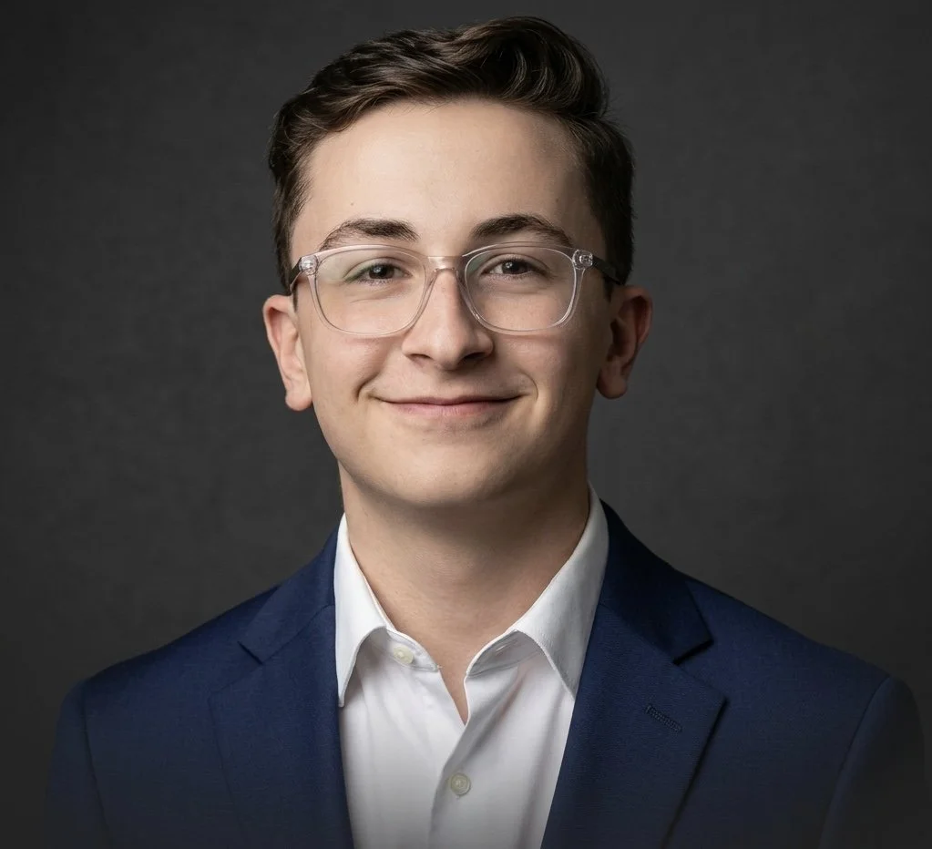 Professional portrait of a young man with glasses, dressed in a suit and white shirt, smiling against a dark gray background.