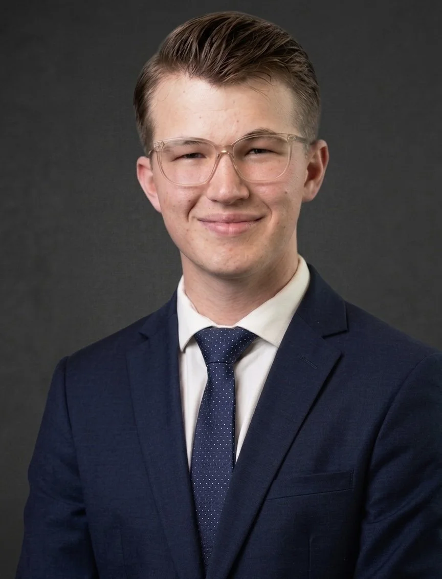Portrait of a young man in a navy suit, white shirt, and blue tie with white polka dots, smiling at the camera, wearing clear glasses, against a dark gray background.
