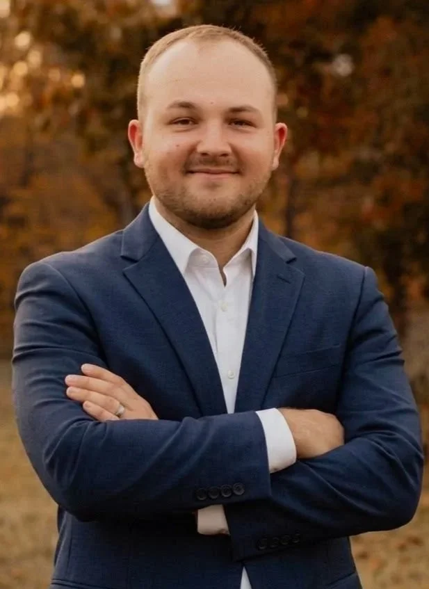 Man in a navy blazer and white shirt with arms crossed, standing outdoors with autumn trees in the background.
