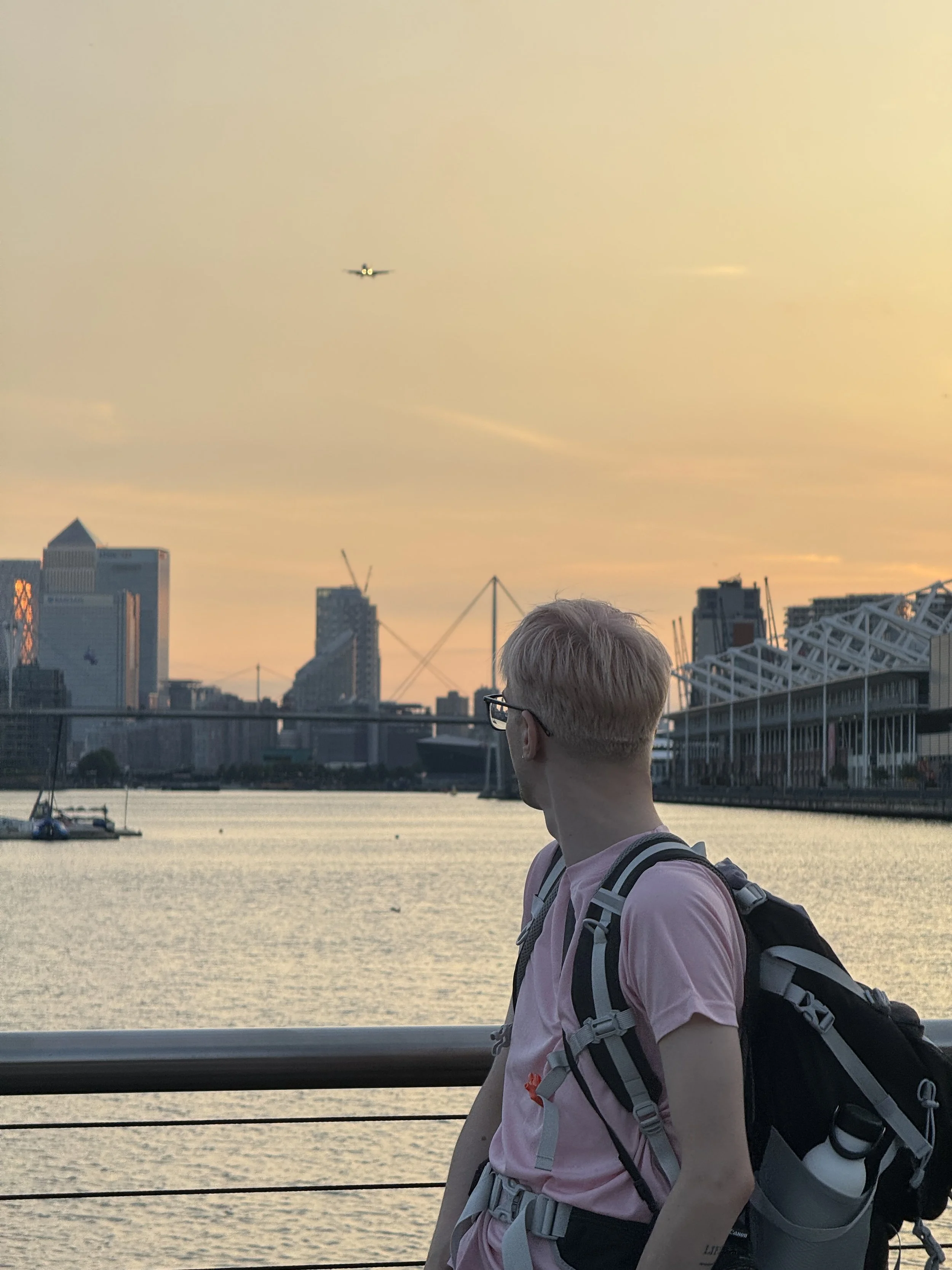 A person with short blonde hair and glasses stands near a body of water, looking away from the camera. In the background, there is a city skyline with high-rise buildings and a boat on the water. An airplane is flying in the sky during sunset.