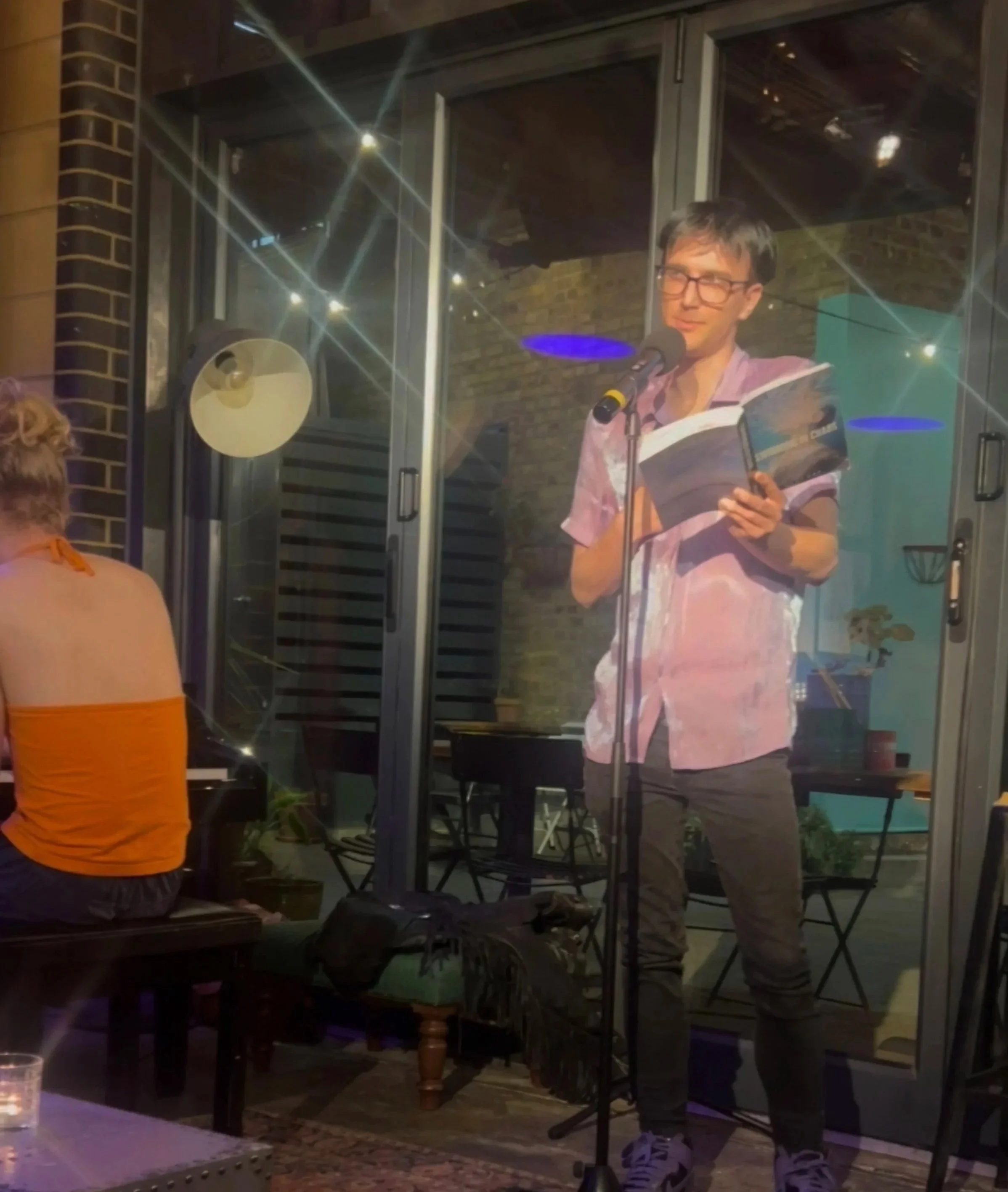 A young man with glasses, wearing a purple shirt, is holding a book and speaking into a microphone indoors at night. The scene can be seen through a glass door, with a woman in an orange top partly visible in the foreground.