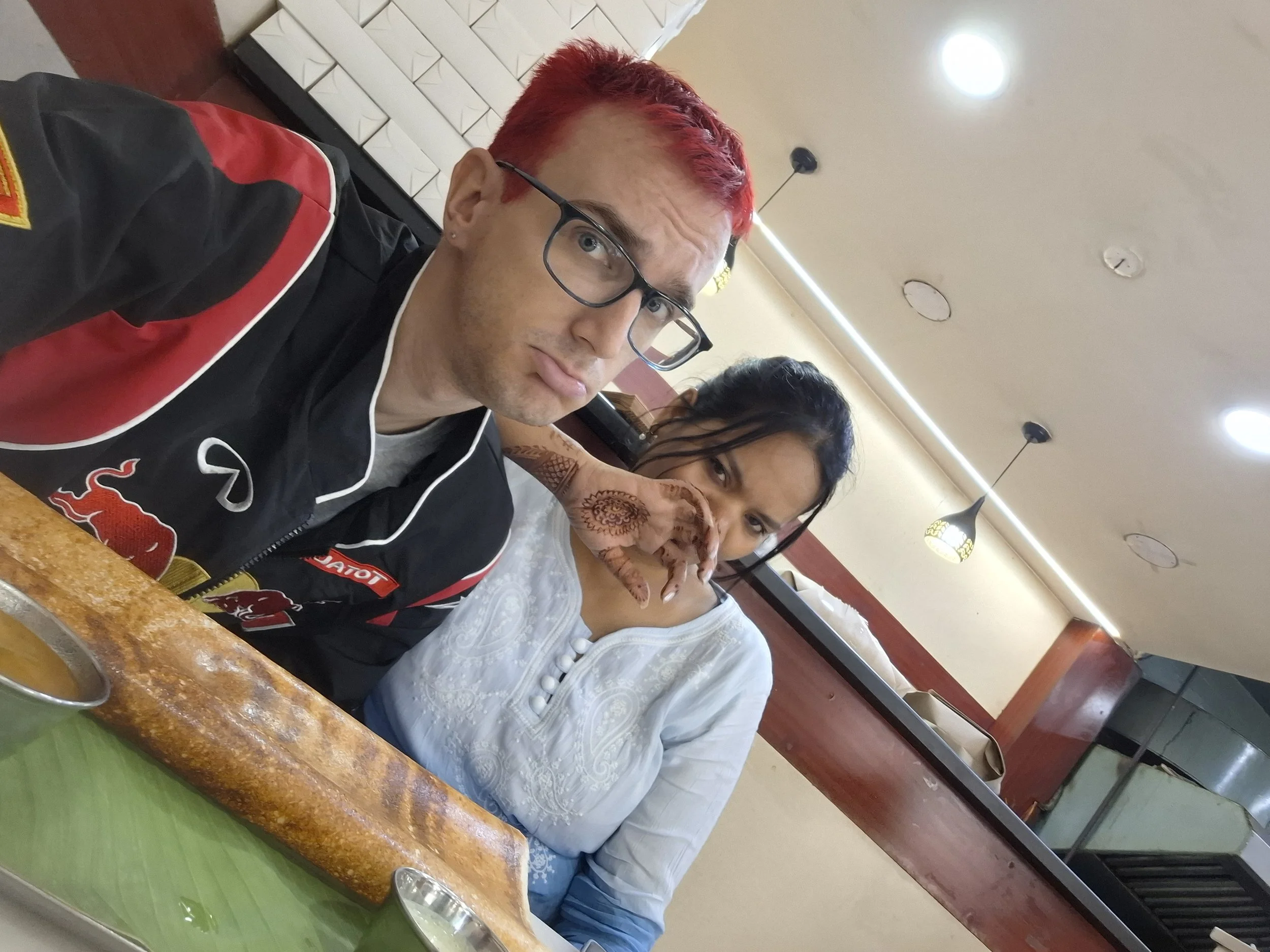 A man with red hair, glasses, and light skin sitting at a restaurant table, with a woman with dark hair and henna on her hand next to him. The man is wearing a black racing jacket with red and white accents. The table has a green cover, and there are hanging lamps and a beige ceiling with circular lights in the background.