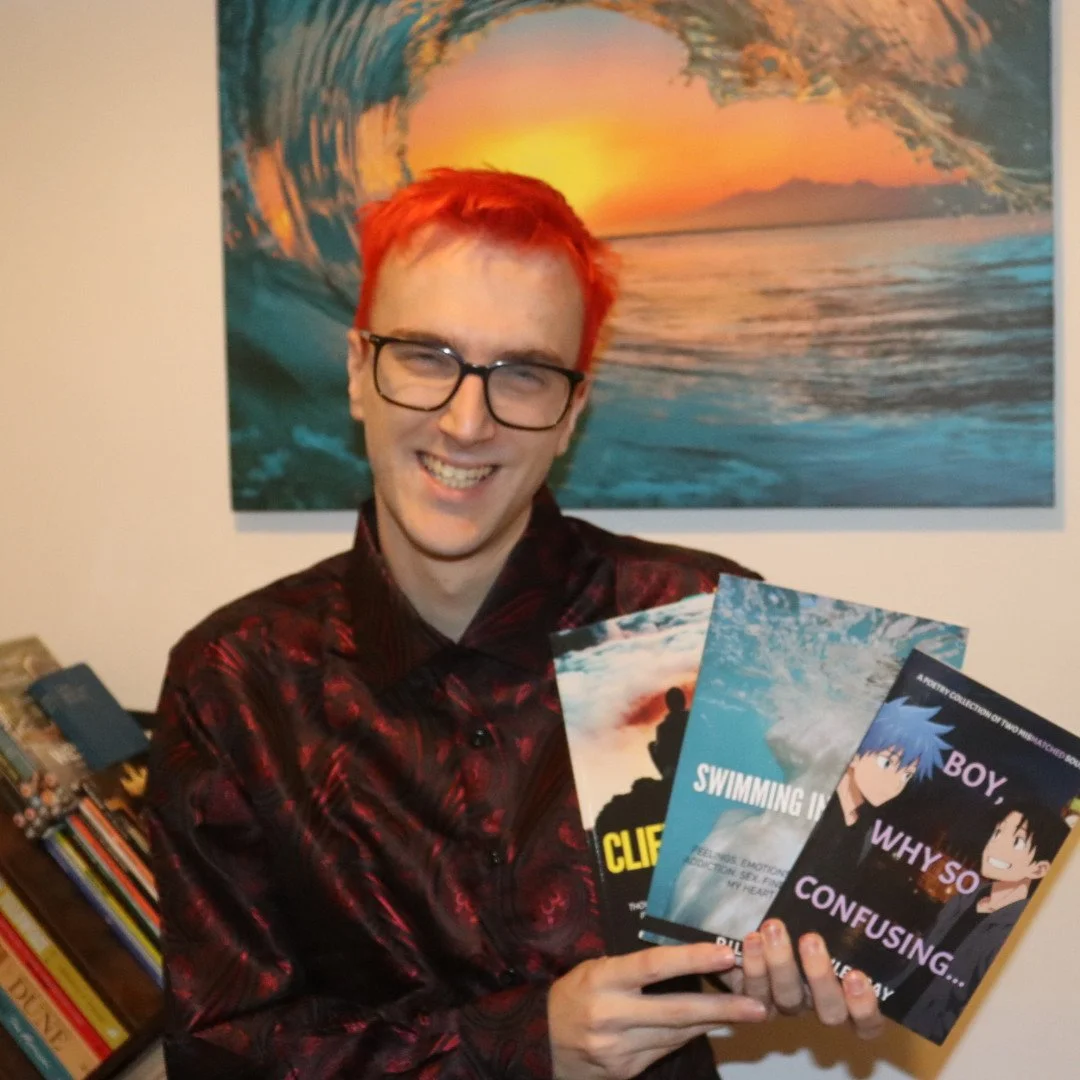 A person with red hair and glasses smiling while holding four books, standing in front of a large wall art of a sunset over the ocean. Books include titles like "Swimming" and "Boy, Why So Confusing." Bookshelf with various books is visible in the background.