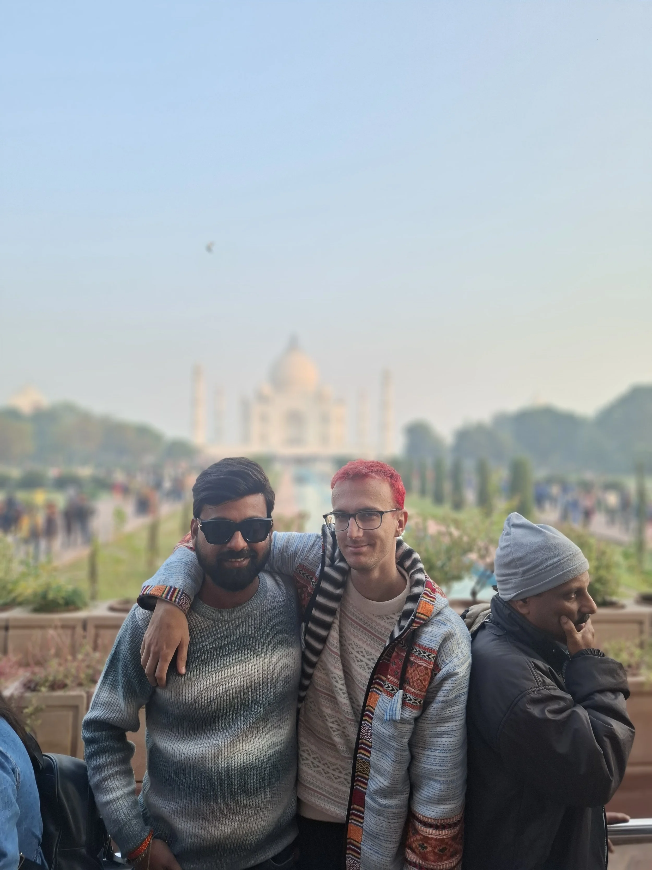 Three friends standing together outdoors with the Taj Mahal in the background, smiling and posing for the photo.