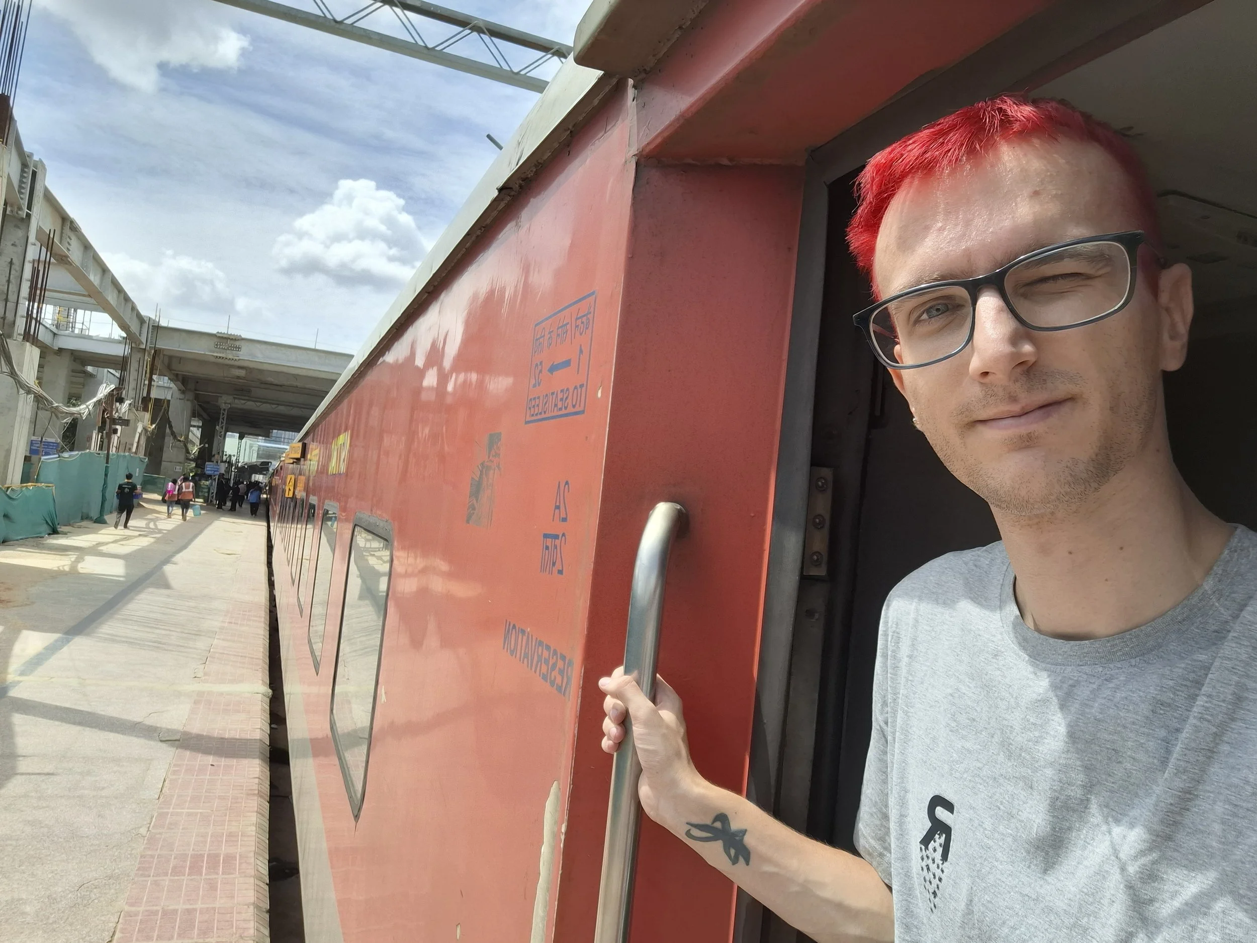 A man with red hair and glasses is leaning out of a red structure, possibly a train, at a busy outdoor station or platform with several people walking in the background under partly cloudy skies.