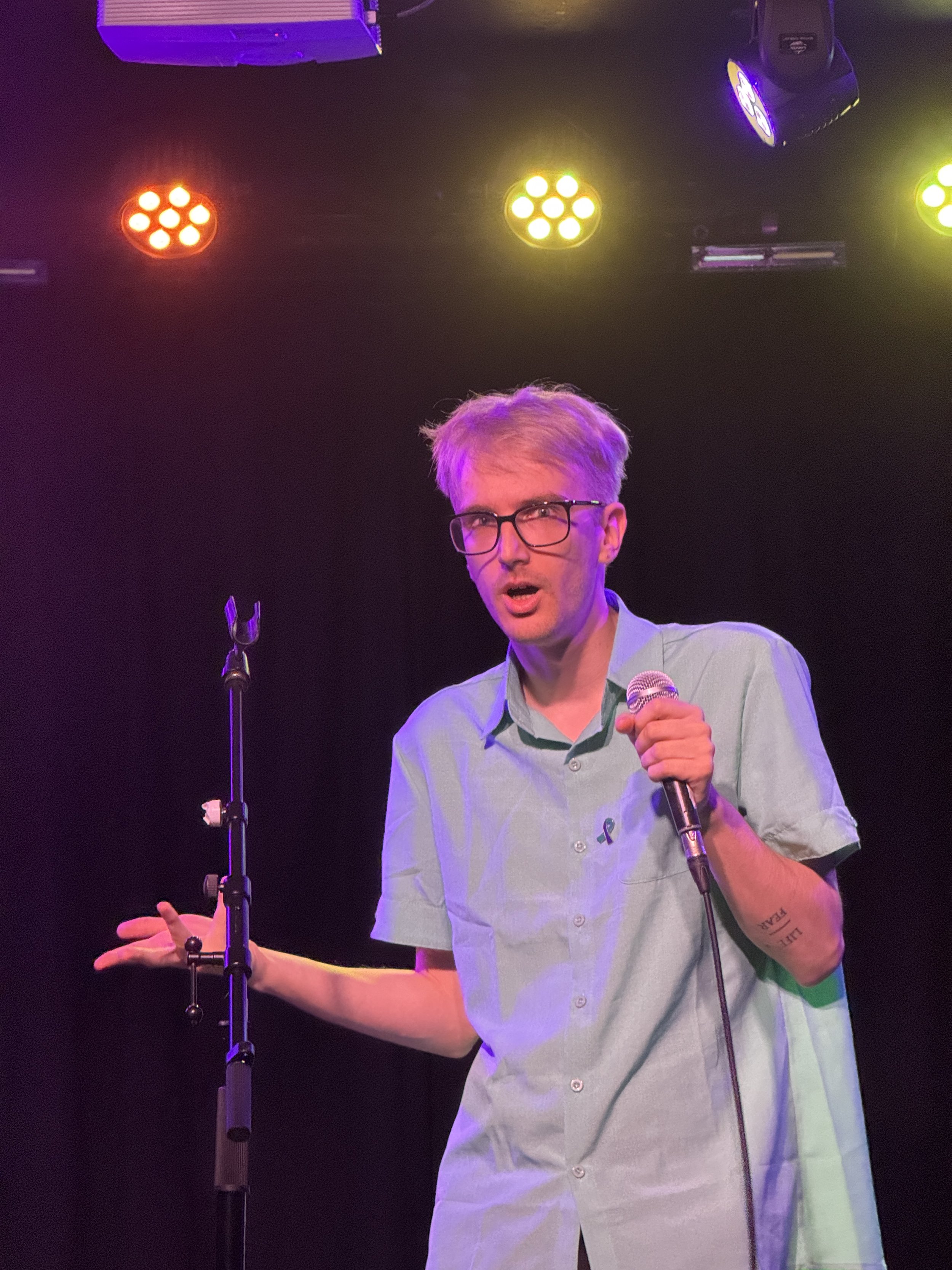 A young man with light hair, glasses, and a green shirt holding a microphone, speaking on stage with colored lights overhead.