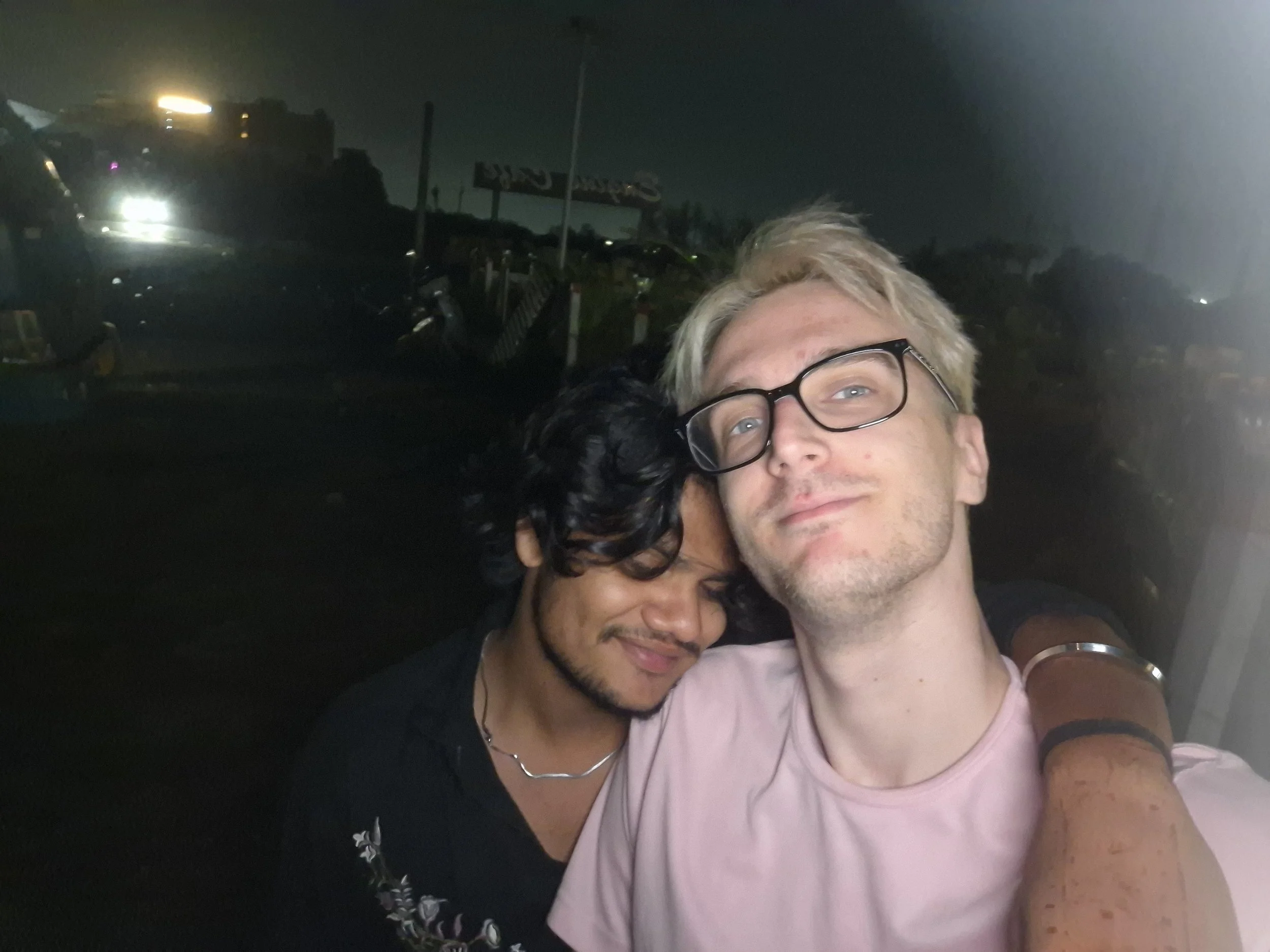 Two people, a woman with dark curly hair and a man with blonde hair and glasses, embracing in a dark outdoor setting at night.