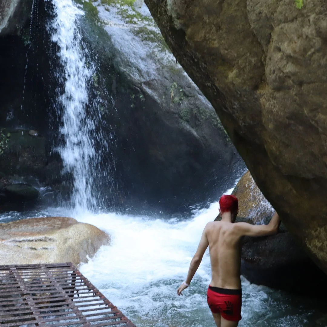 A person with red hair wearing red shorts standing under a small waterfall in a rocky canyon.