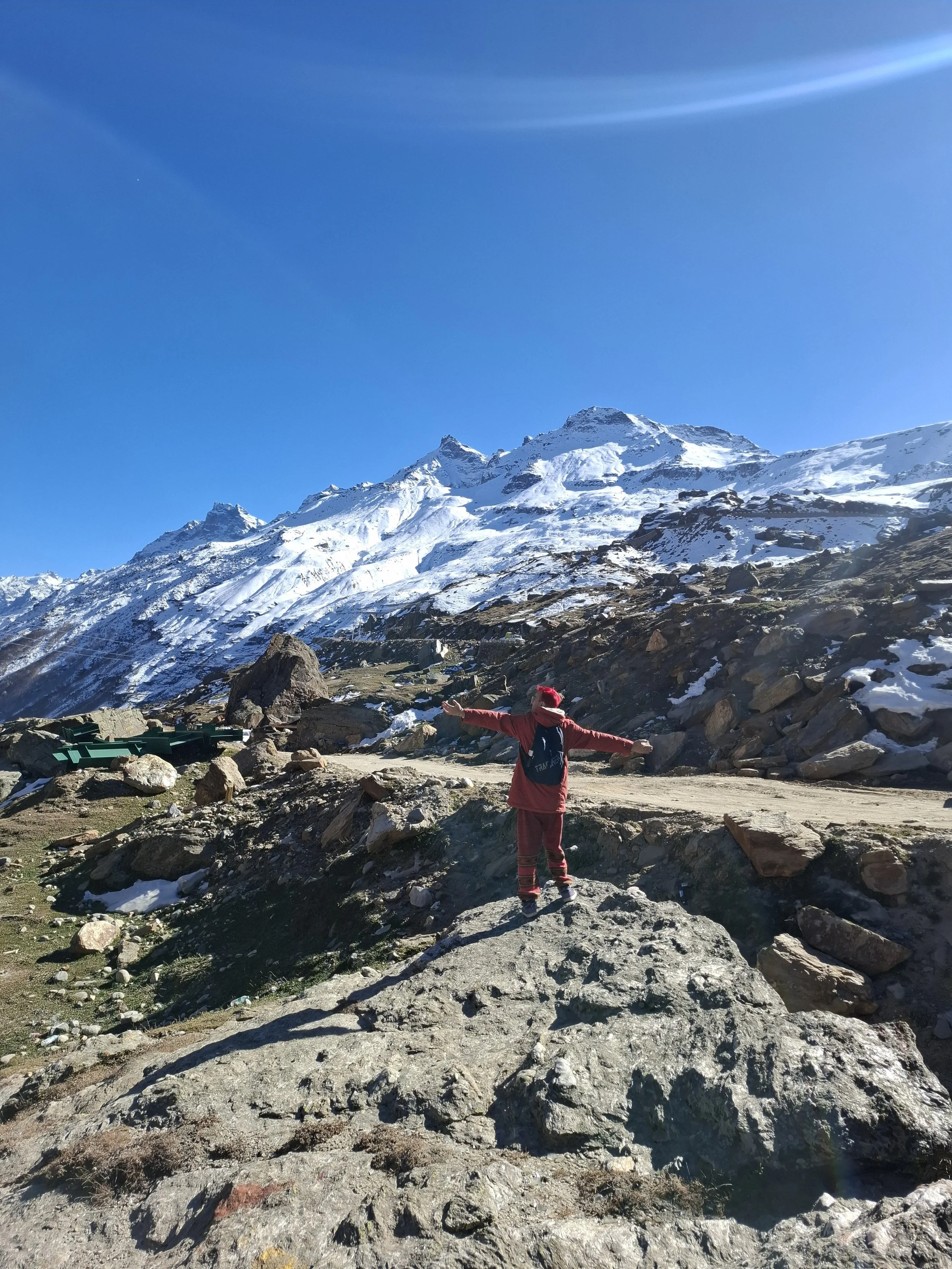 Person wearing red jacket with arms outstretched standing on rocky terrain in front of snow-covered mountain peaks under a clear blue sky.
