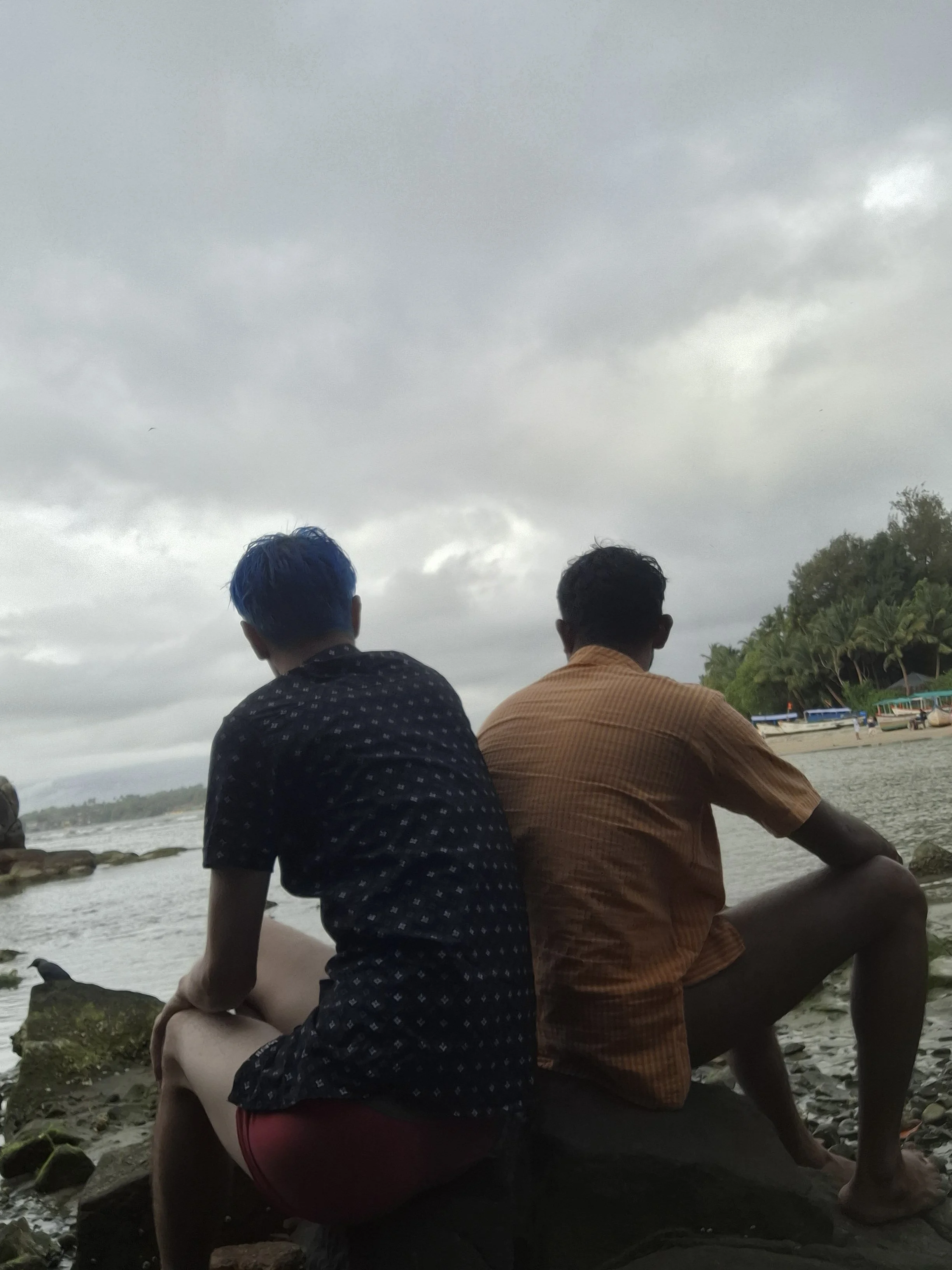 Two men sitting by the water on a rocky shore, facing away from the camera, with a cloudy sky overhead and boats on the beach in the distance.