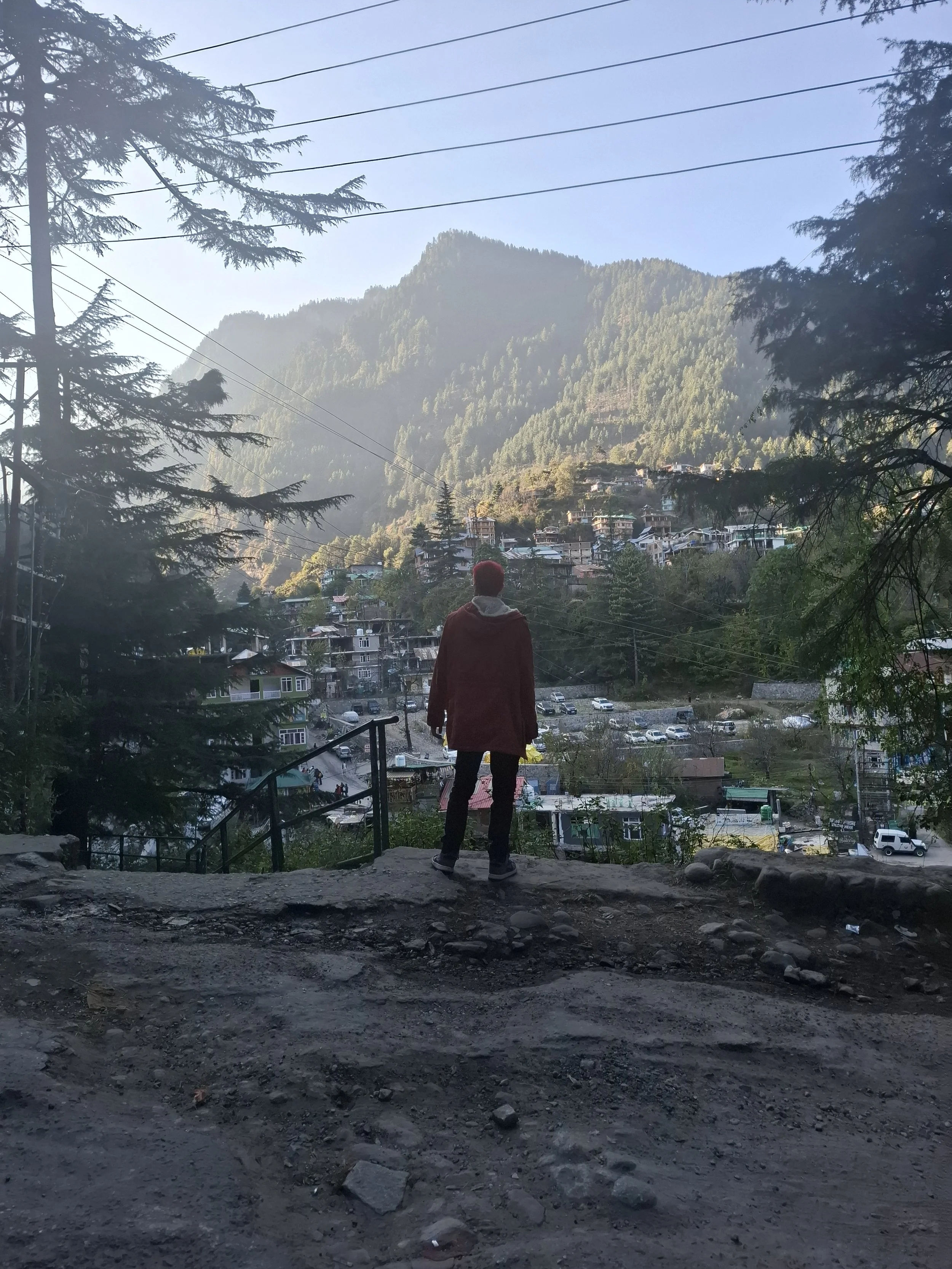 Person standing on a stone path overlooking a town in a valley with houses, surrounded by mountains and trees.