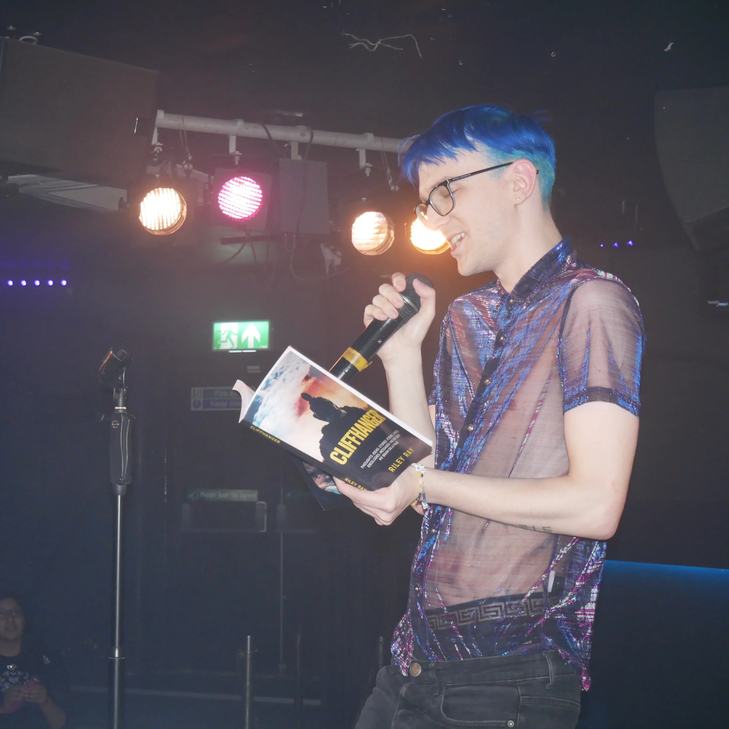 A young person with short, blue hair holding a microphone and a booklet, standing in front of a stage with colorful lights and a dark background.