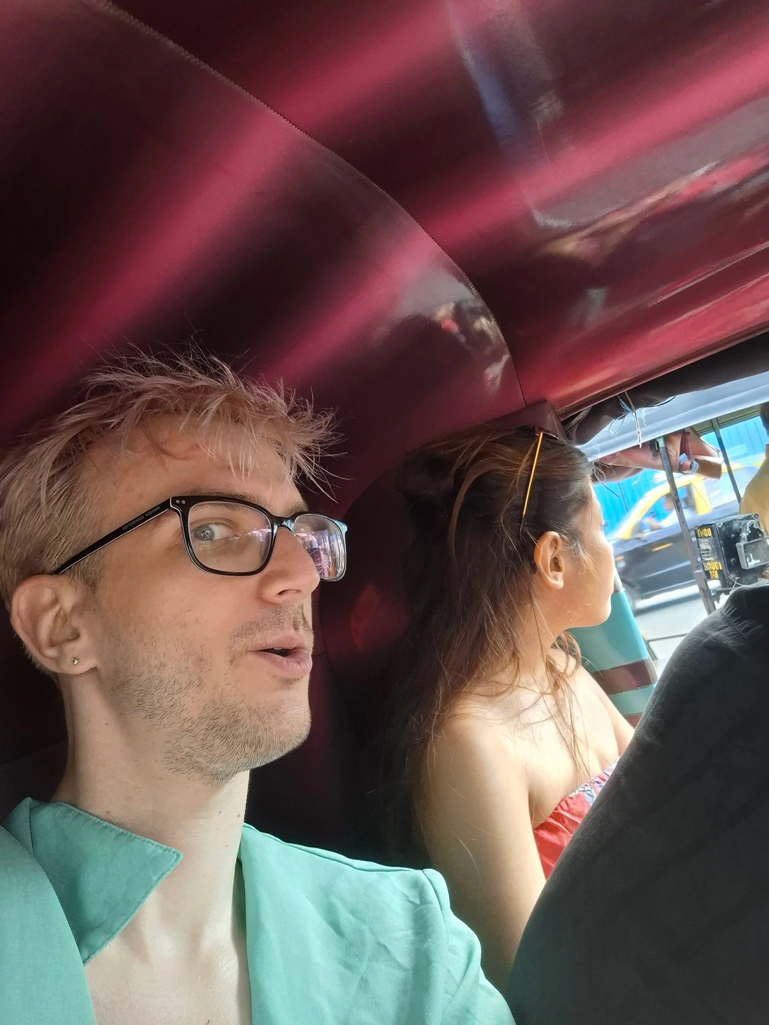 Two people sitting in a vehicle, one man with glasses and a woman with brown hair, both facing forward, visible from the shoulder up.