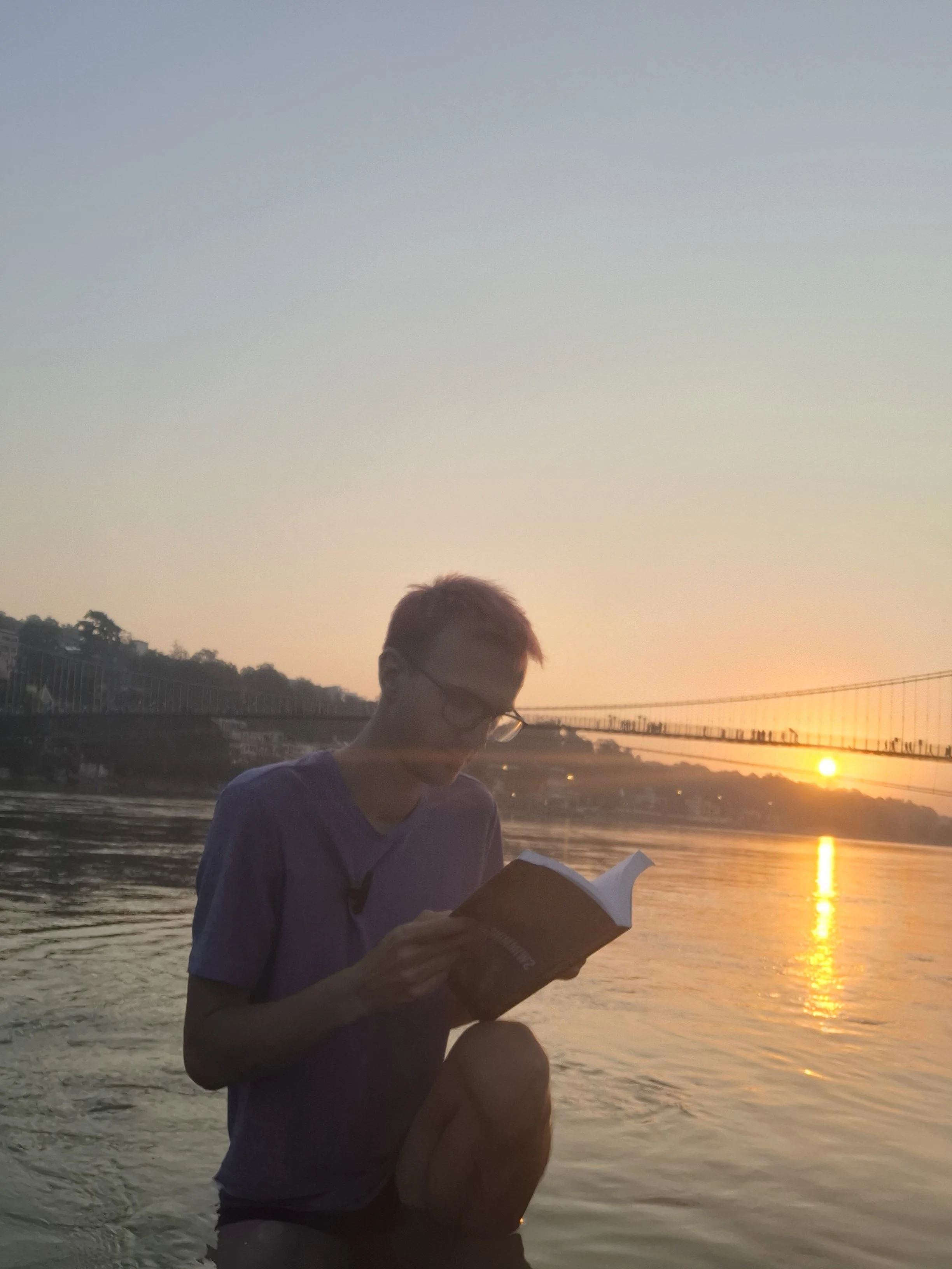 A person with glasses reading a book by the water at sunset with a bridge and trees in the background.