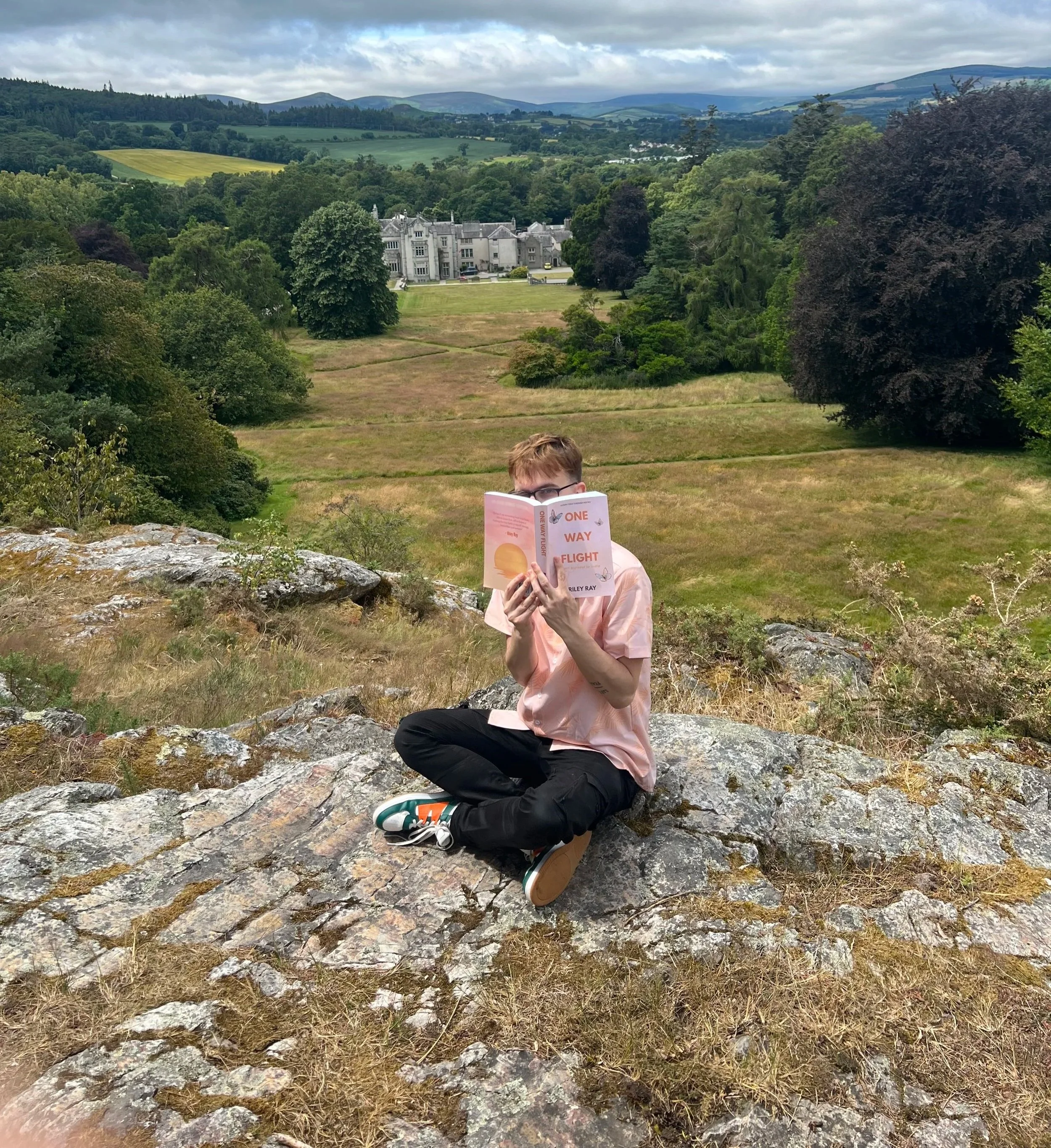 A person with short brown hair and glasses is sitting on rocky ground, reading a book titled 'One Way Flight' in an outdoor setting with grass, trees, and distant rolling hills under a cloudy sky.