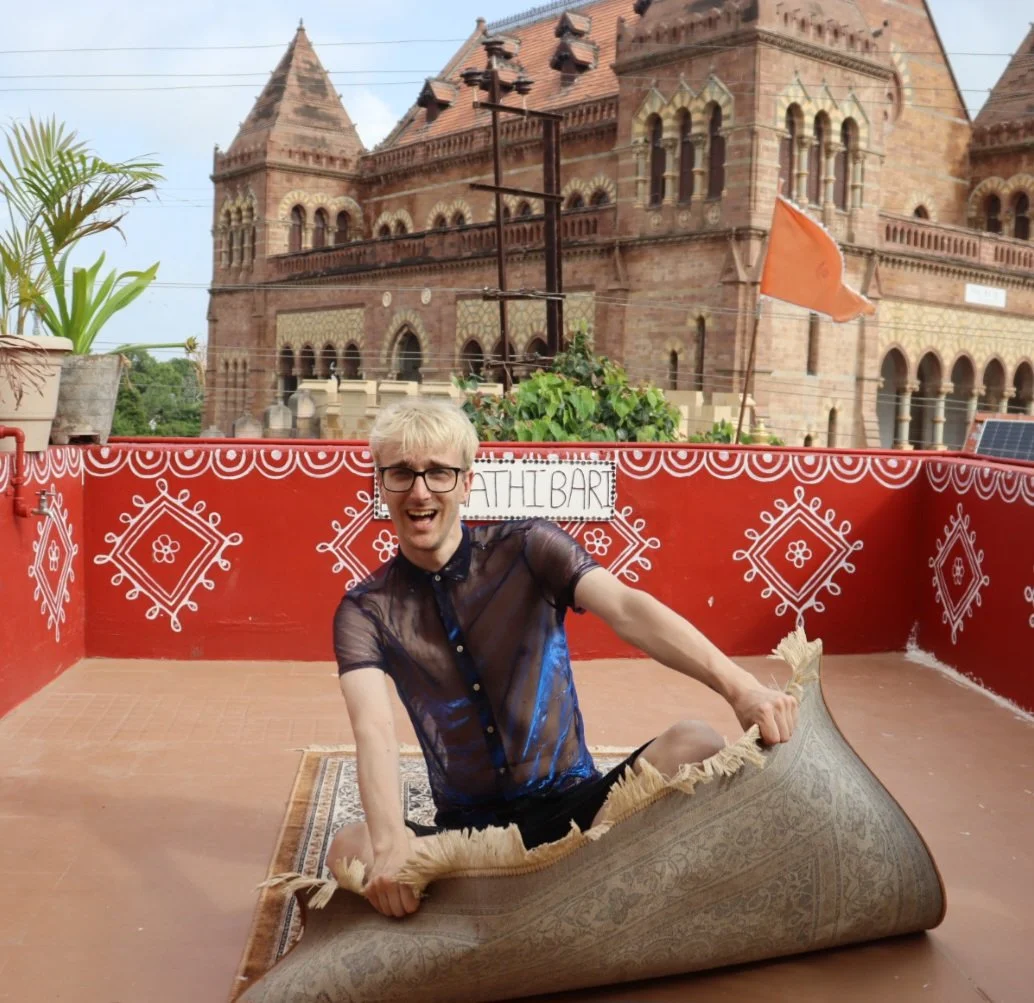 A person with blonde hair, glasses, and a sheer black shirt sitting on a rolled-up rug on a terrace with red patterned walls. Behind them is a historic brick building and a sign that says 'ATH BARI'.