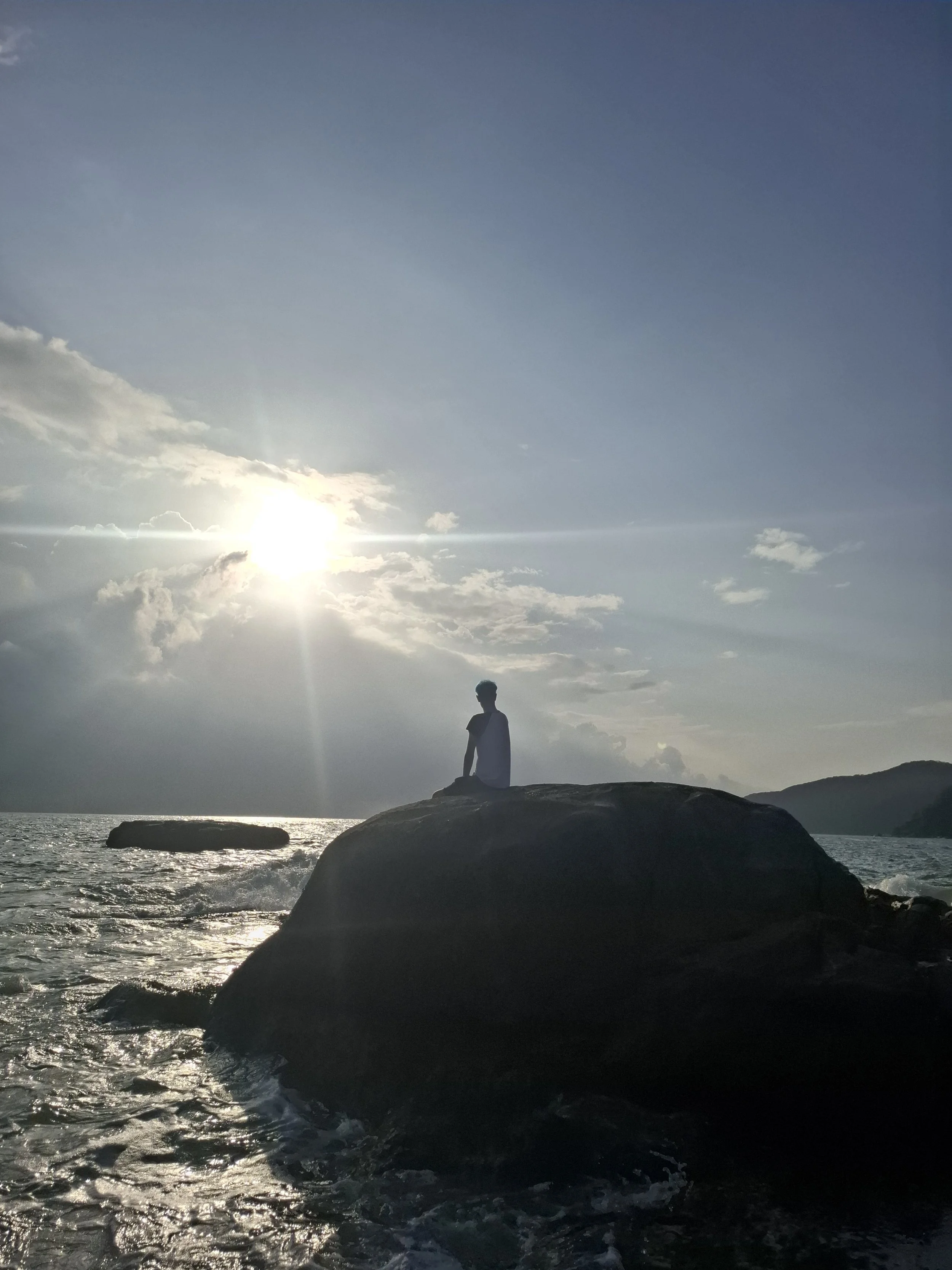 A silhouette of a person sitting on a large rock by the ocean with the sun shining through clouds in the sky.