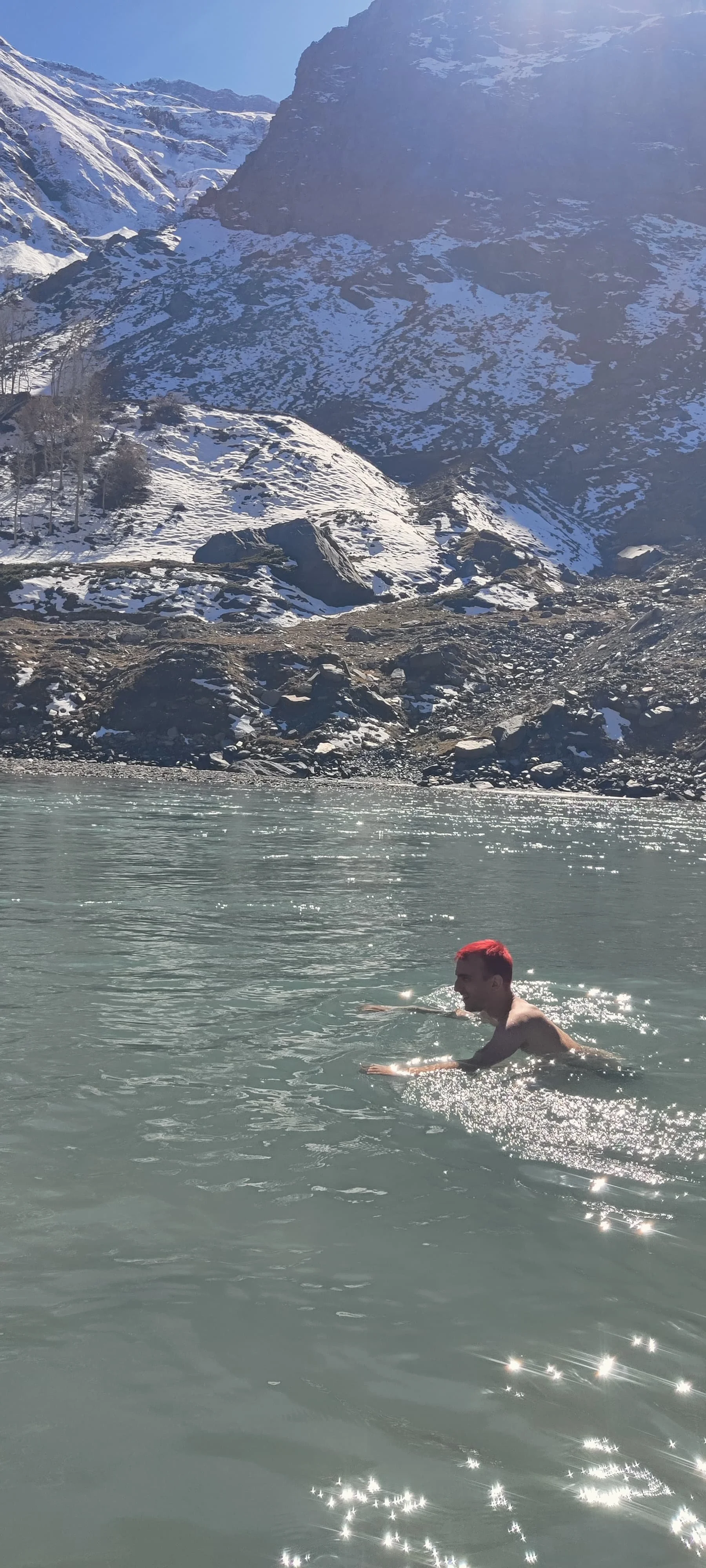 A person with red hair swimming in a mountain lake with snowy peaks and rocky terrain in the background.