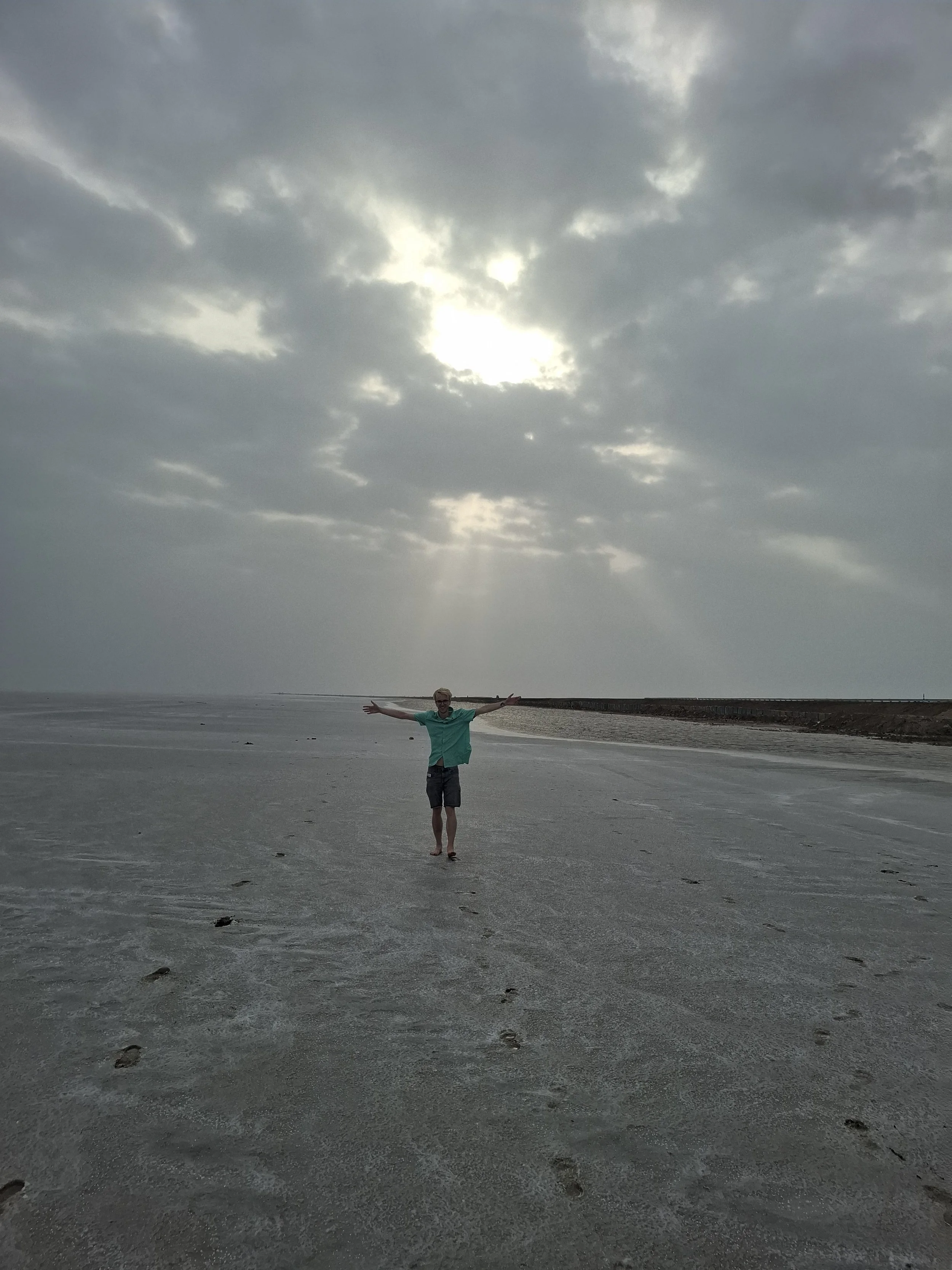 A person standing barefoot on a sandy beach with arms outstretched under a cloudy sky with the sun barely visible through the clouds.