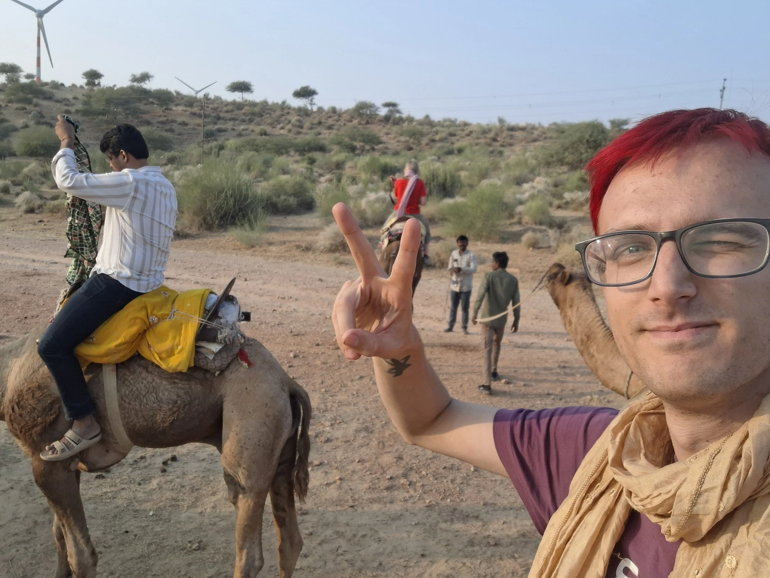 A person with red hair and glasses making a peace sign while riding a camel in a desert area with sparse vegetation and wind turbines in the background.