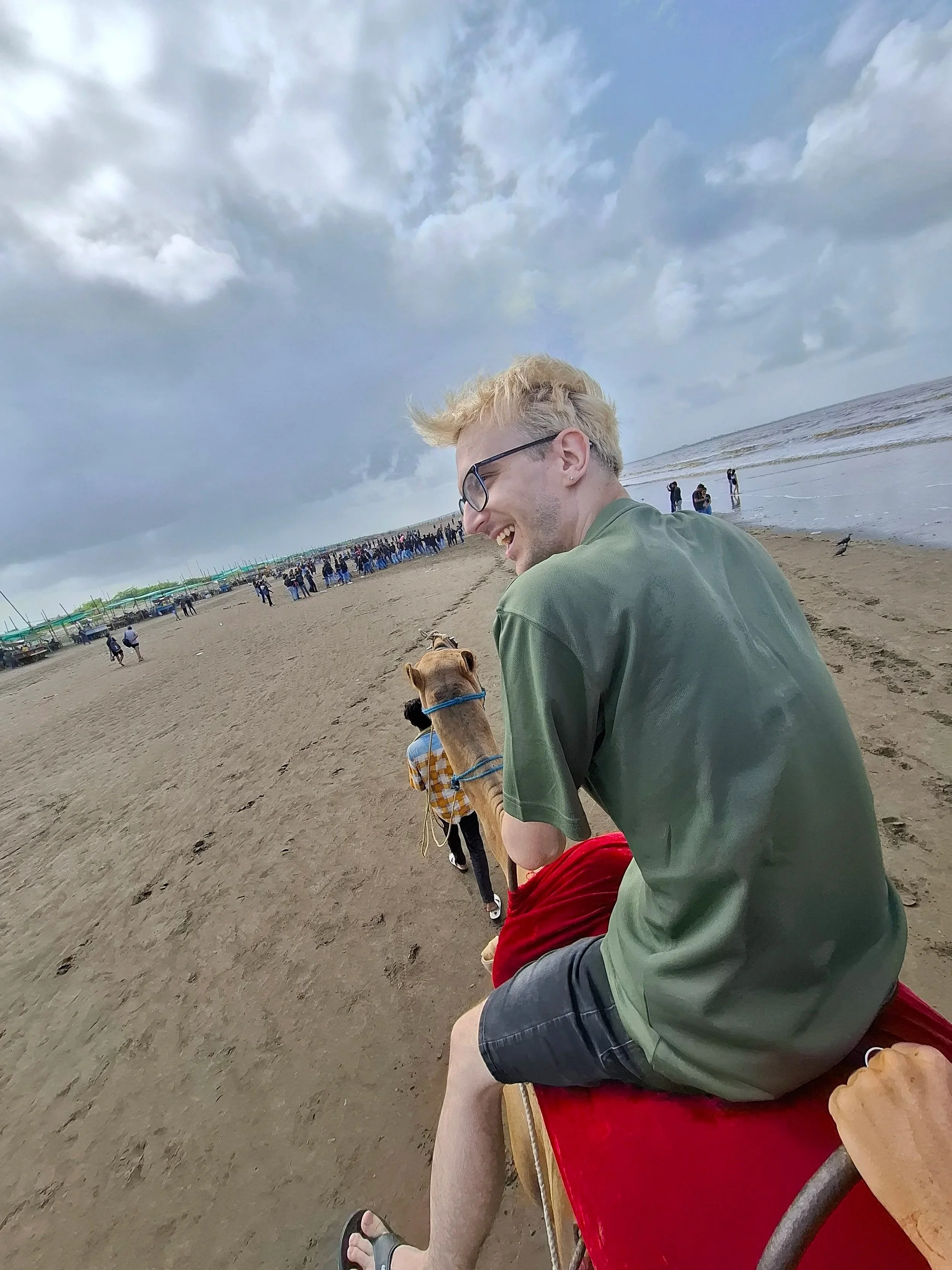 A young man with blonde hair, glasses, and a green shirt laughing on a beach, sitting next to a camel with a person in the background, cloudy sky, and ocean waves.