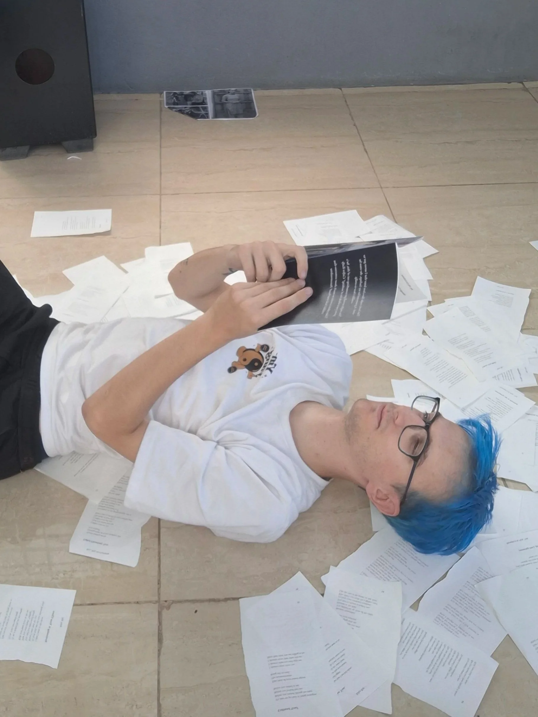 Person with blue hair lying on the floor surrounded by scattered papers, holding a booklet.