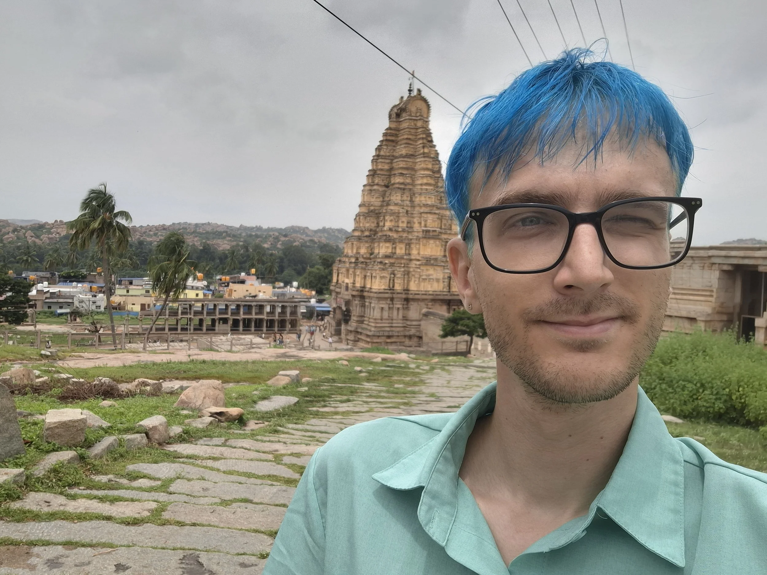 A young man with glasses and blue hair taking a selfie at an ancient temple or monument with a pagoda-like tower in the background, surrounded by greenery and a cloudy sky.