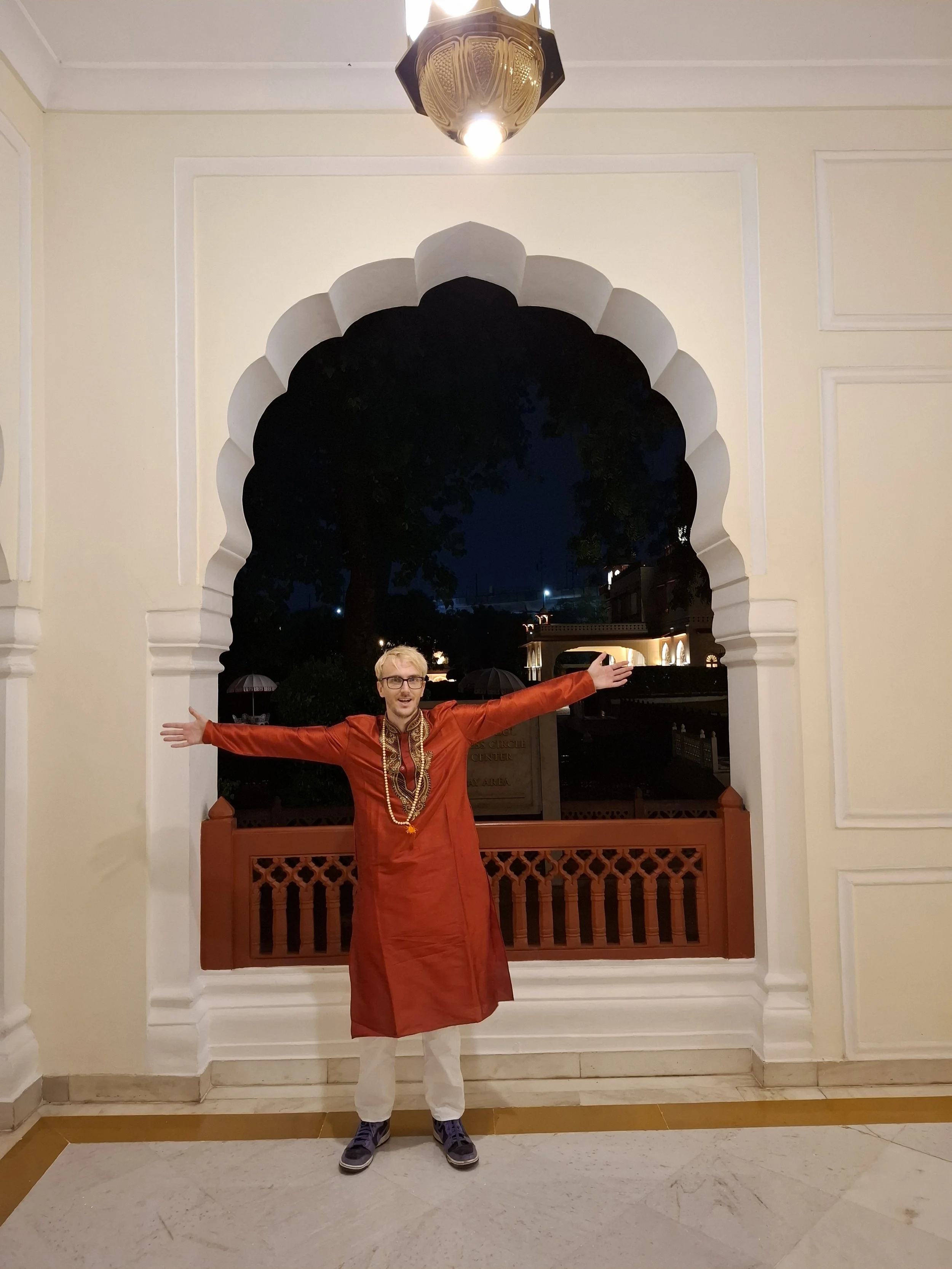 A man dressed in traditional Indian attire with arms outstretched standing under an ornate archway at night.