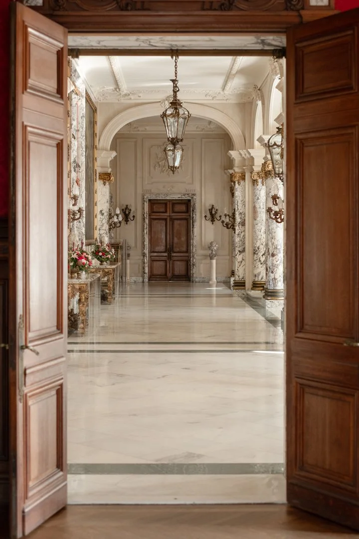 View of an elegant, ornate hallway with white marble floors and architectural details, seen through a wooden doorway.