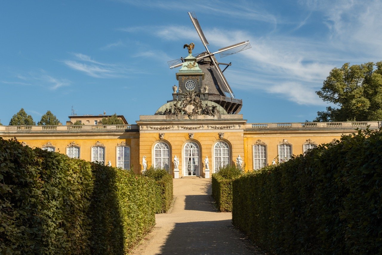 A historic building with classical architecture, featuring large arched windows, white statues outside, and a yellow facade, topped with a traditional windmill.