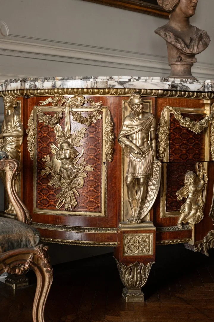 An ornate wooden cabinet with gold decorative sculptures of figures and garlands on its front, topped with a marble slab, in a room with classical art busts and sculptures.