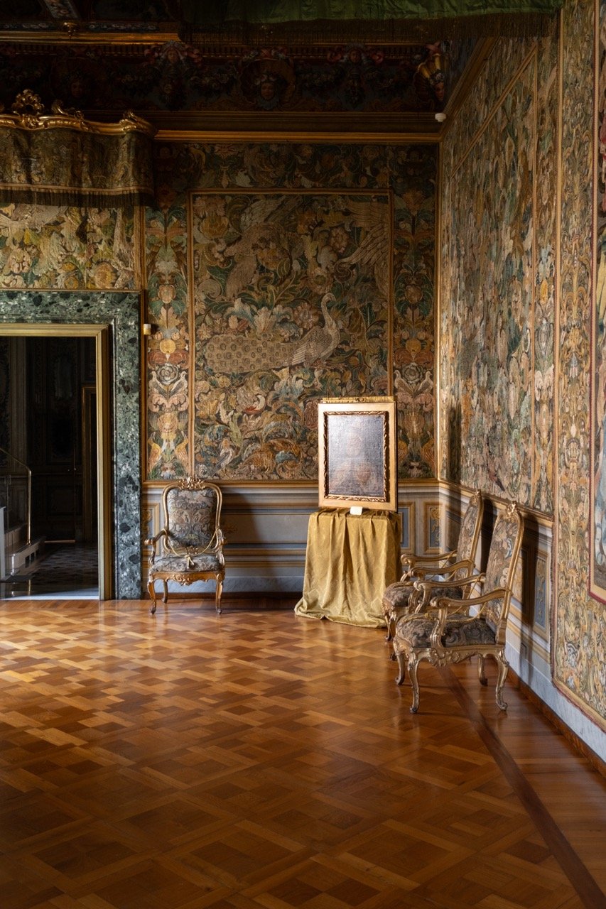 Interior of an ornate room with wood parquet flooring, decorated wallpaper, and antique chairs. A framed artwork is displayed on a covered pedestal, with floral and animal motifs on the walls.