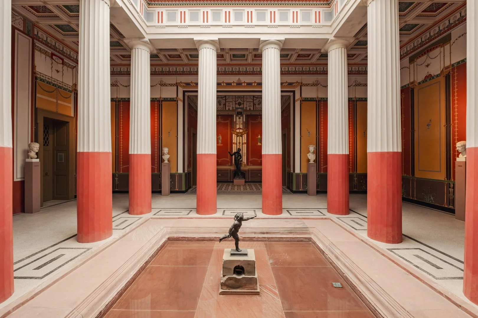 Interior view of a classical building with tall, white fluted columns with red bases, ornate painted walls, and marble flooring. There are bust sculptures on pedestals along the walls and statues in the background. In the foreground, there is a small