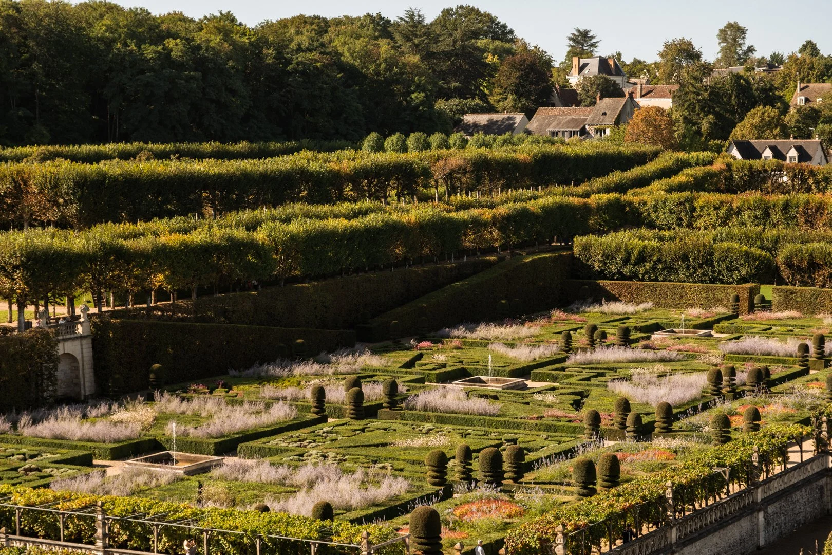 A formal garden with neatly trimmed hedges, topiary, and small fountains, surrounded by lush greenery and houses in the background.