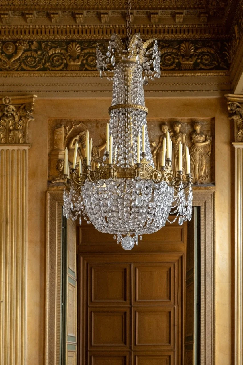 Elegant crystal chandelier hanging from ornate gold ceiling with wood-paneled doors and classical sculptures in the background.