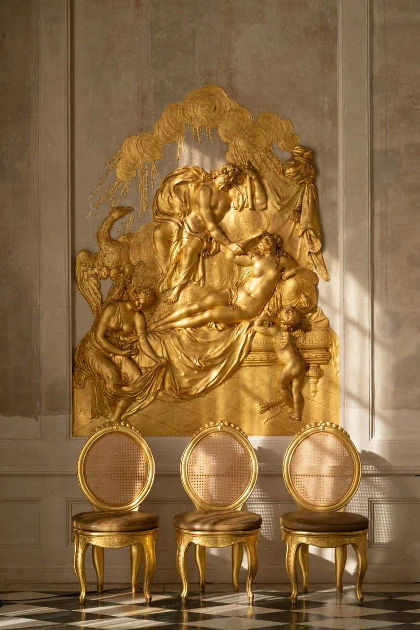 A large, ornate gold wall sculpture depicting a classical scene with multiple figures, including a reclining woman and standing figures, with a background of clouds and drapery. Three gold chairs with oval backs and cushioned seats are in front of th