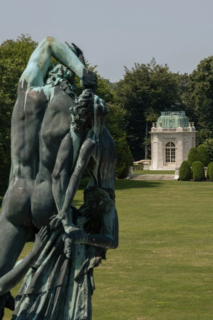 A bronze sculpture of multiple intertwined figures in a park with a historic building and lush trees in the background.