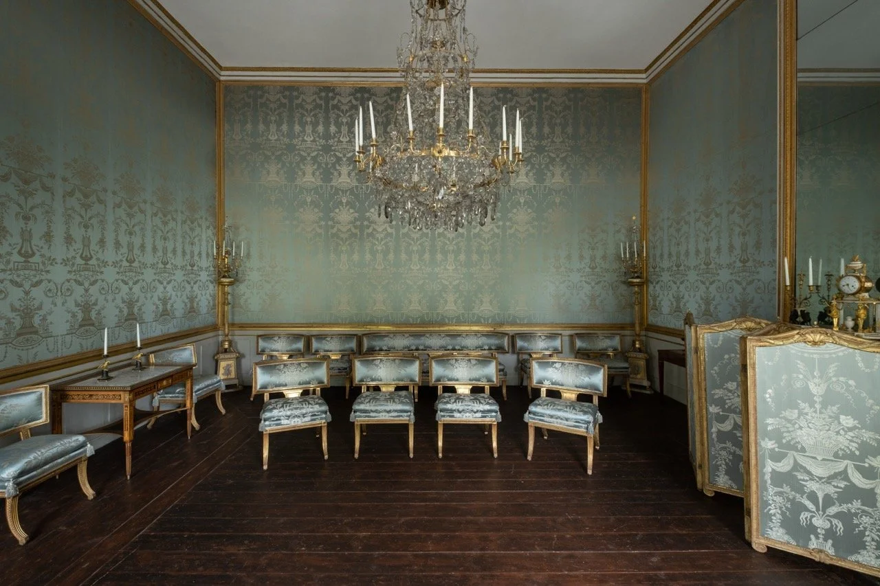 Elegant room with ornate blue and gold wallpaper, a large crystal chandelier, gold-trimmed chairs, a wooden table with candelabras, and a dark wood floor.