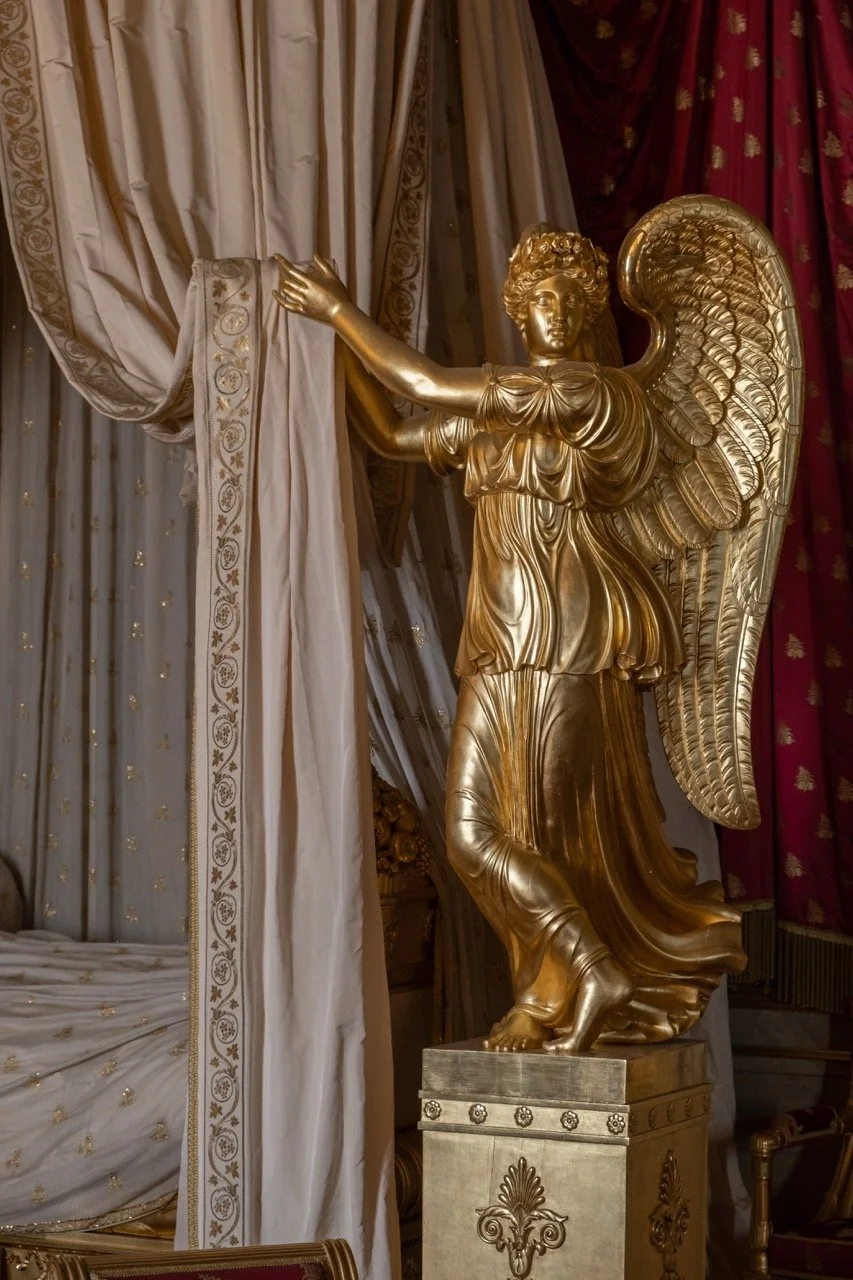 A large gold-colored statue of a winged figure with curly hair, wearing flowing robes, with one arm extended forward, standing on a decorated pedestal inside an ornate room with gold and red curtains.