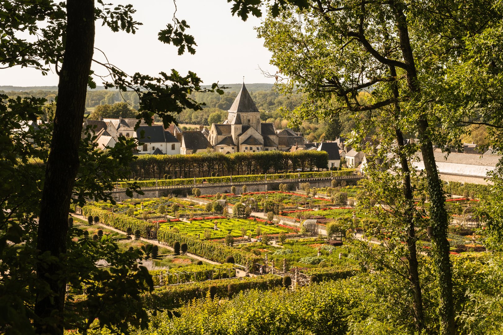 Scenic view of a European village with a church and well-maintained gardens, framed by trees.