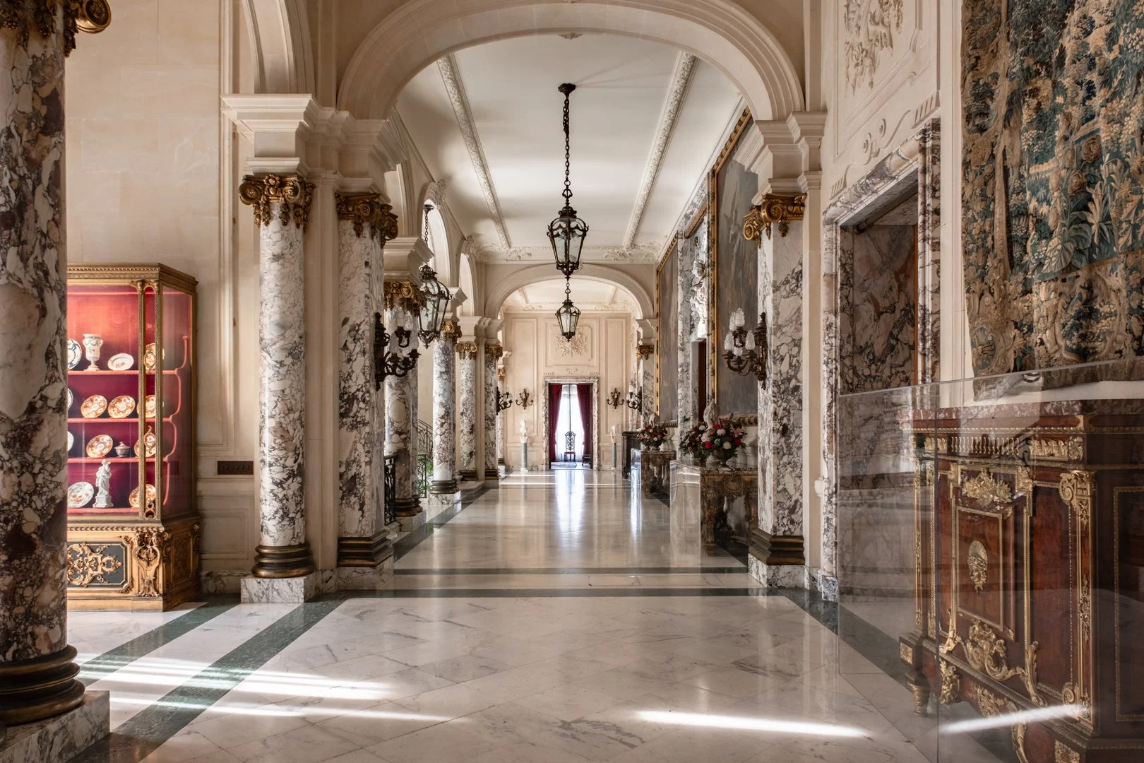 Elegant marble hallway with ornate columns, chandeliers, and floral arrangements, leading to a doorway with red curtains.