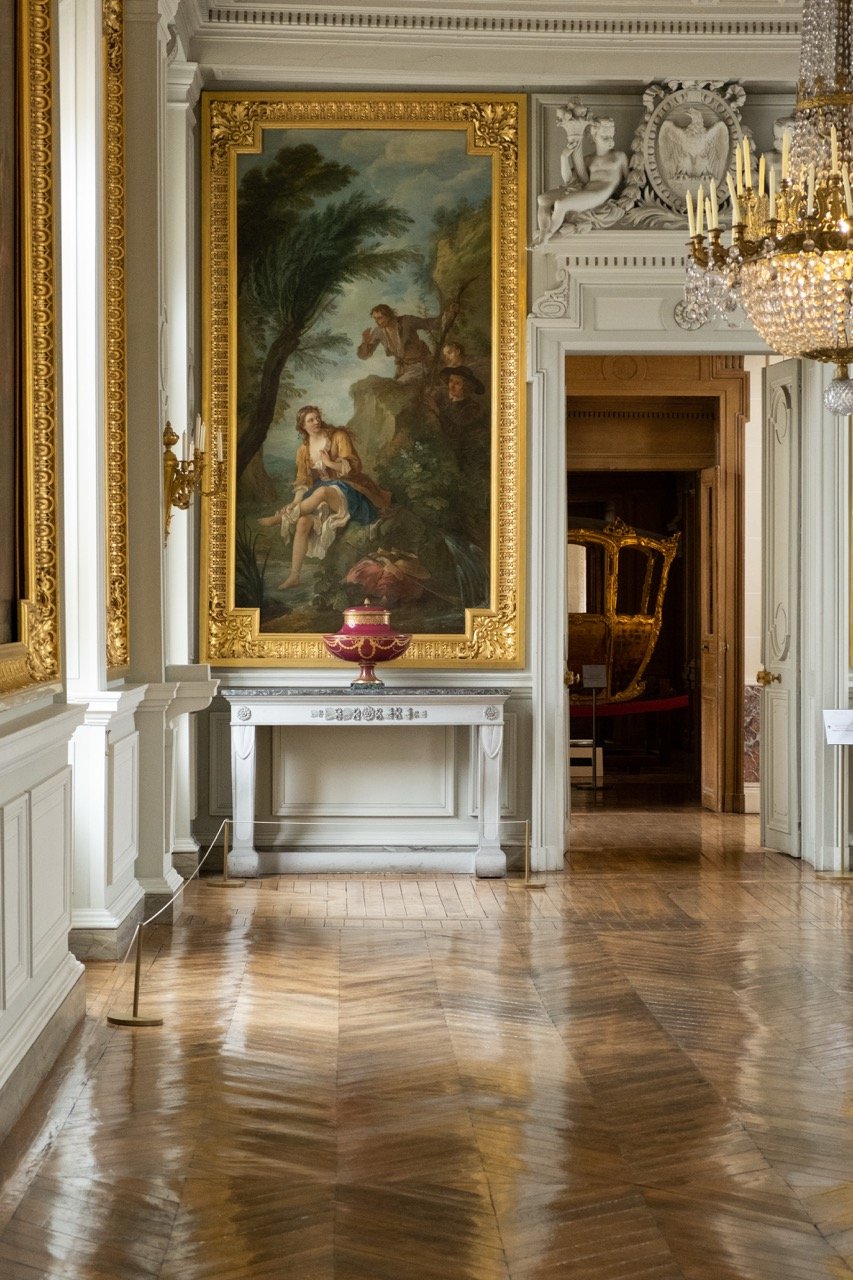 Elegant room with large framed oil painting on wall, ornate gold frame, white and gold decor, chandelier, wood flooring, decorative sculptures, and a red decorative vase on a white table.